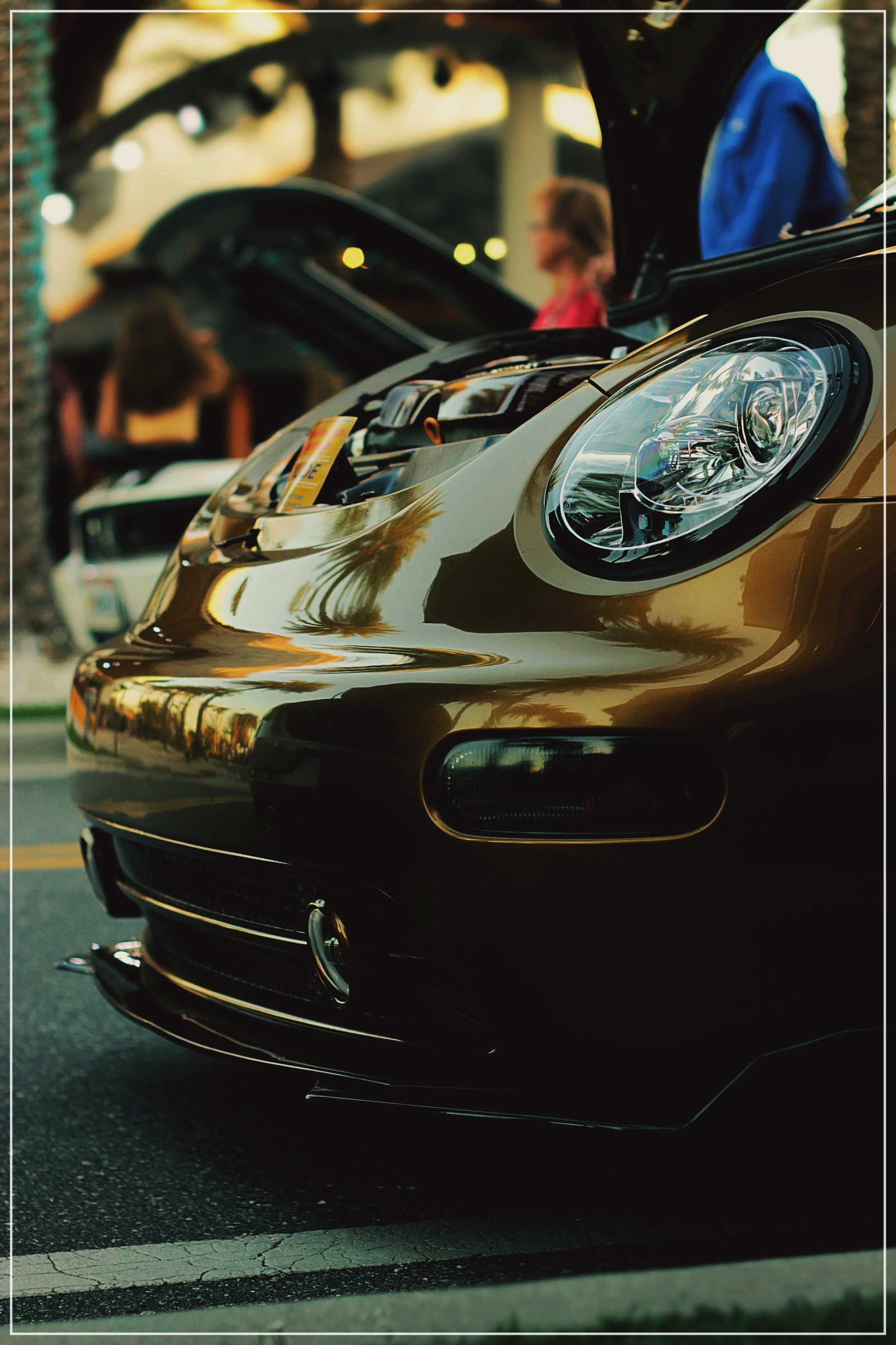 Close-up of a shiny gold sports car with its hood open, parked at an outdoor car show, with other vehicles and people in the background.