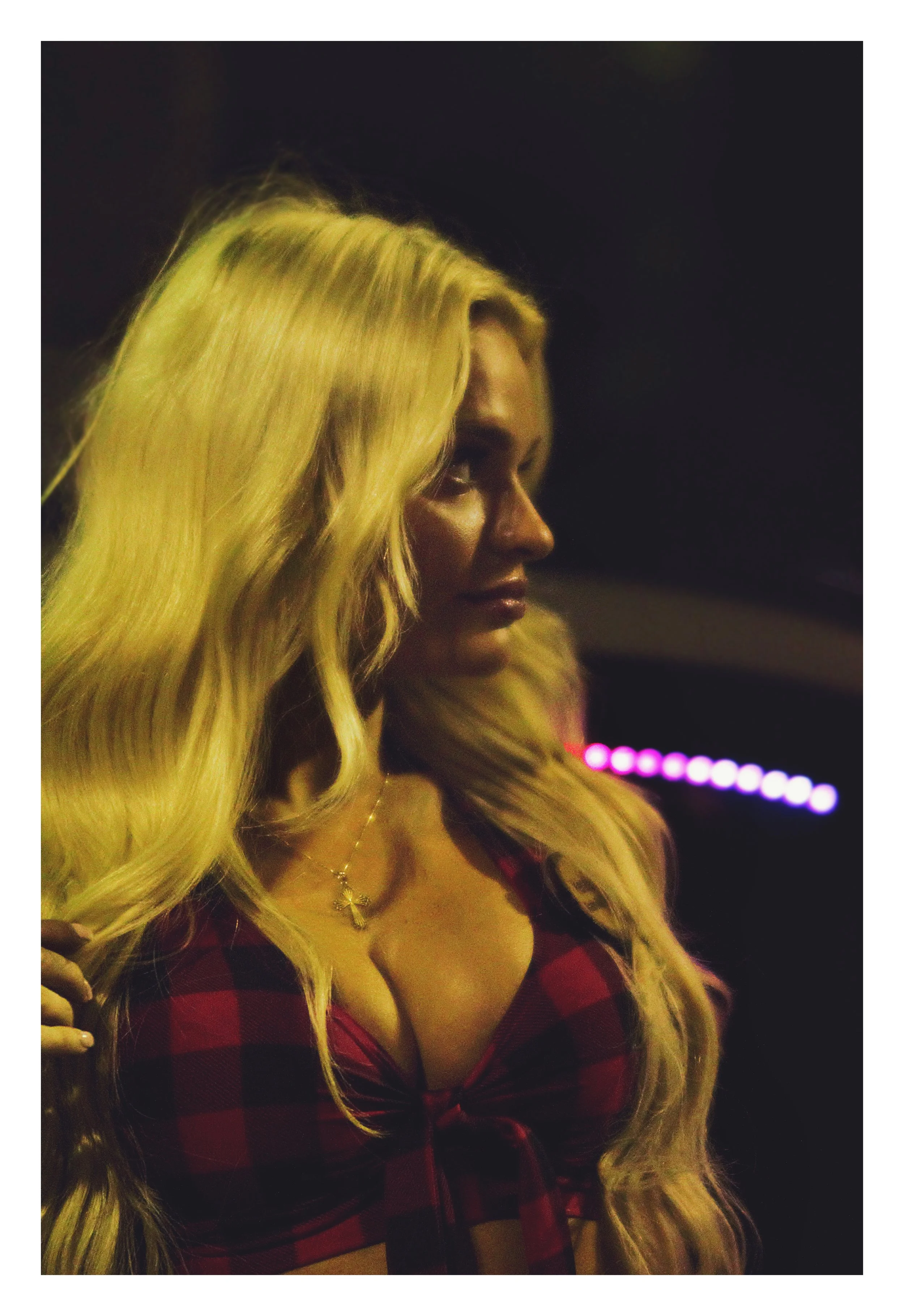 A woman with long, wavy blonde hair wearing a red and black plaid shirt, looking to her left with a serious expression, in a dimly lit setting with colorful lights in the background.