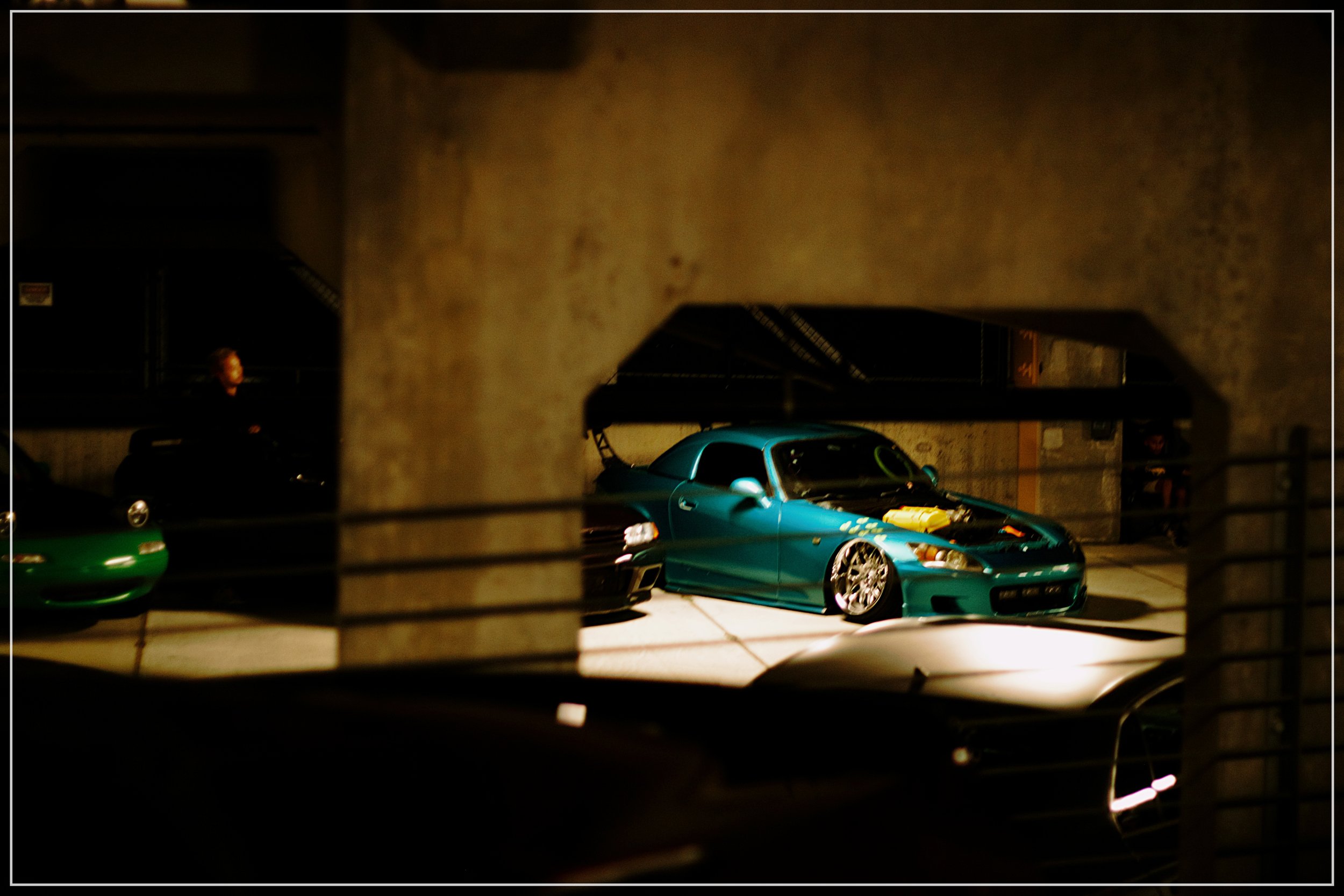 A teal-colored modified sports car parked in a dimly lit indoor parking garage, viewed through a concrete barrier with metal railings.