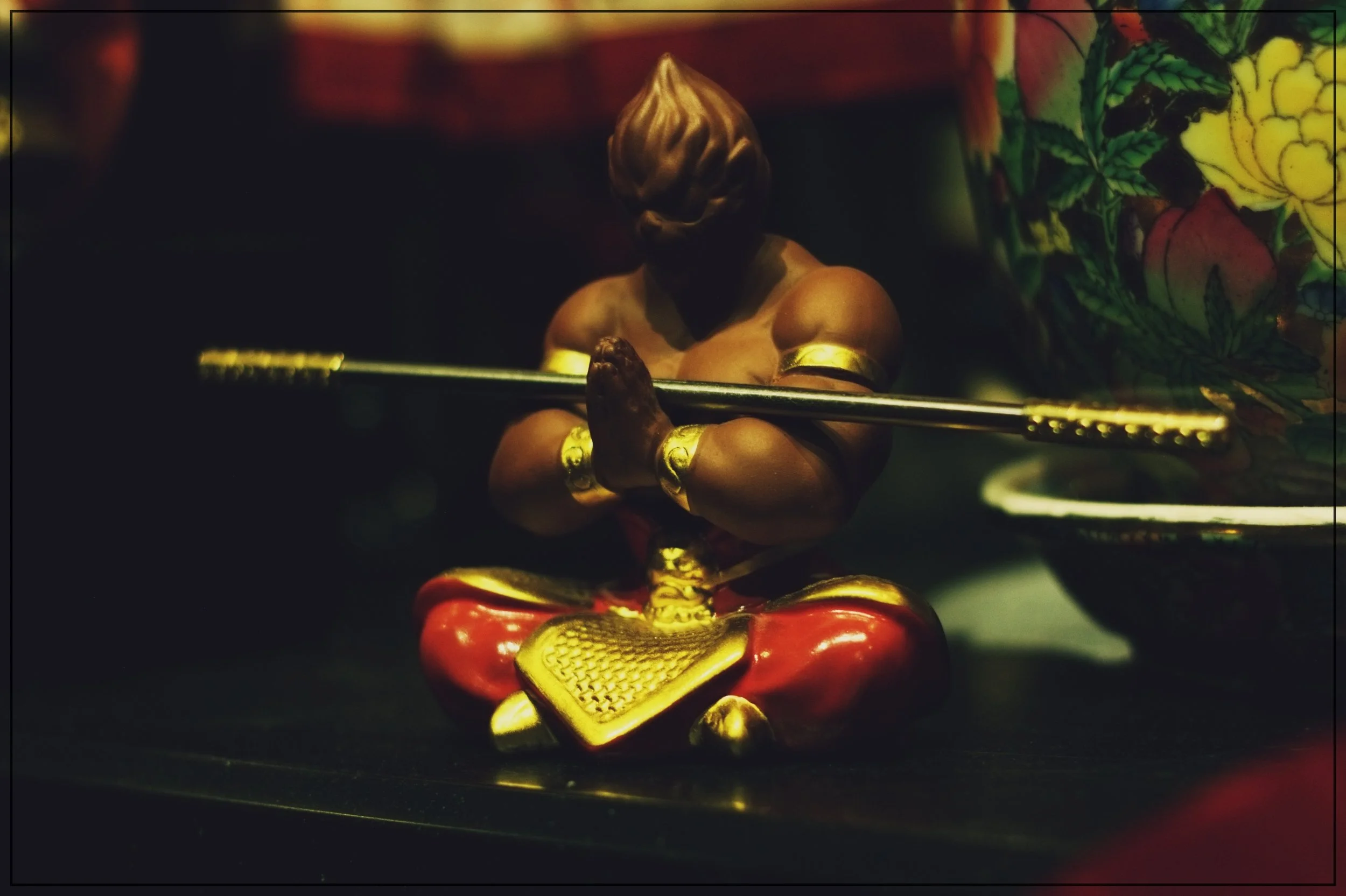 A statue of a muscular person with a dark face, holding a spear, seated with crossed legs, dressed in red and gold clothing with decorative gold accessories, positioned on a black surface in low lighting.