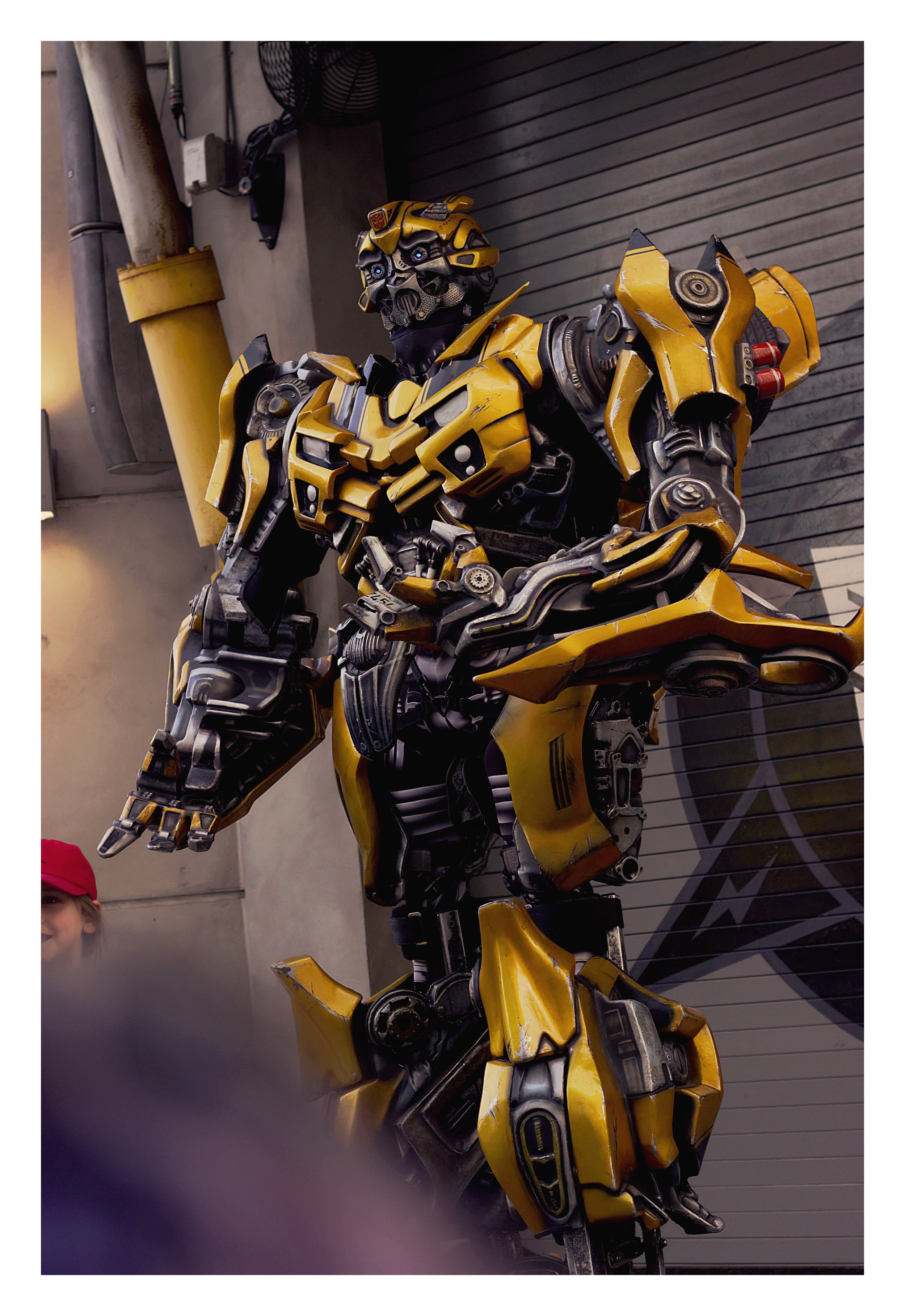 A life-sized yellow Transformers character, Bumblebee, showcasing robotic features and detailed mechanical parts, positioned against a gray wall with garage-style door.