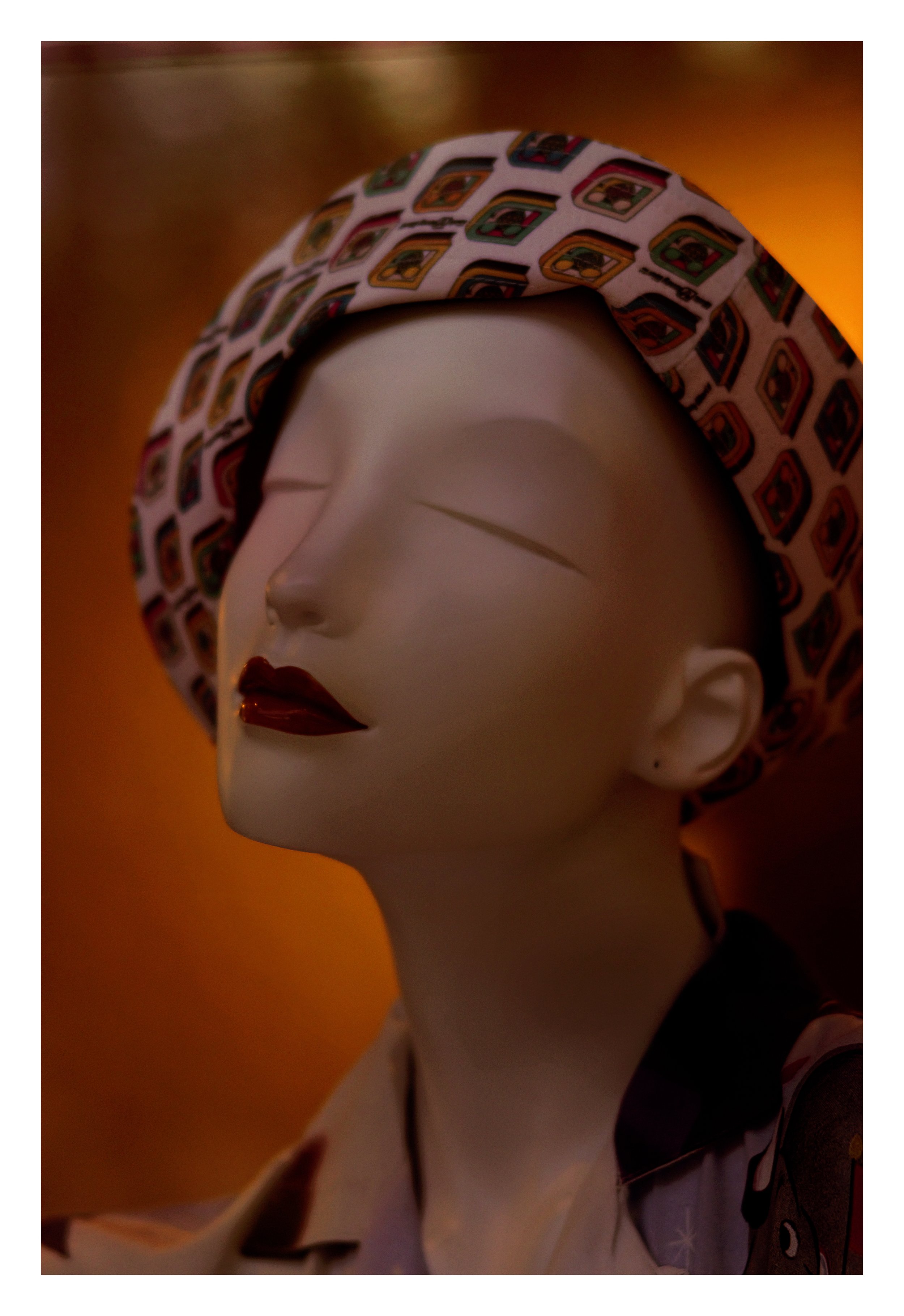 A mannequin with a glossy face, closed eyes, dark red lipstick, and a colorful hat with a pattern of small images, set against a warm, gradient background.
