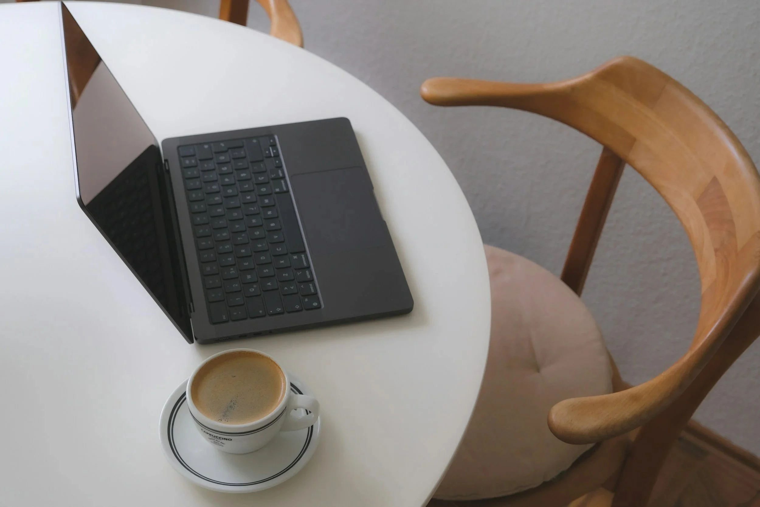 A white round table with a black laptop and a cup of coffee on a saucer. A wooden chair with a beige cushion is partially visible. Quiet space ready for telehealth sessions.