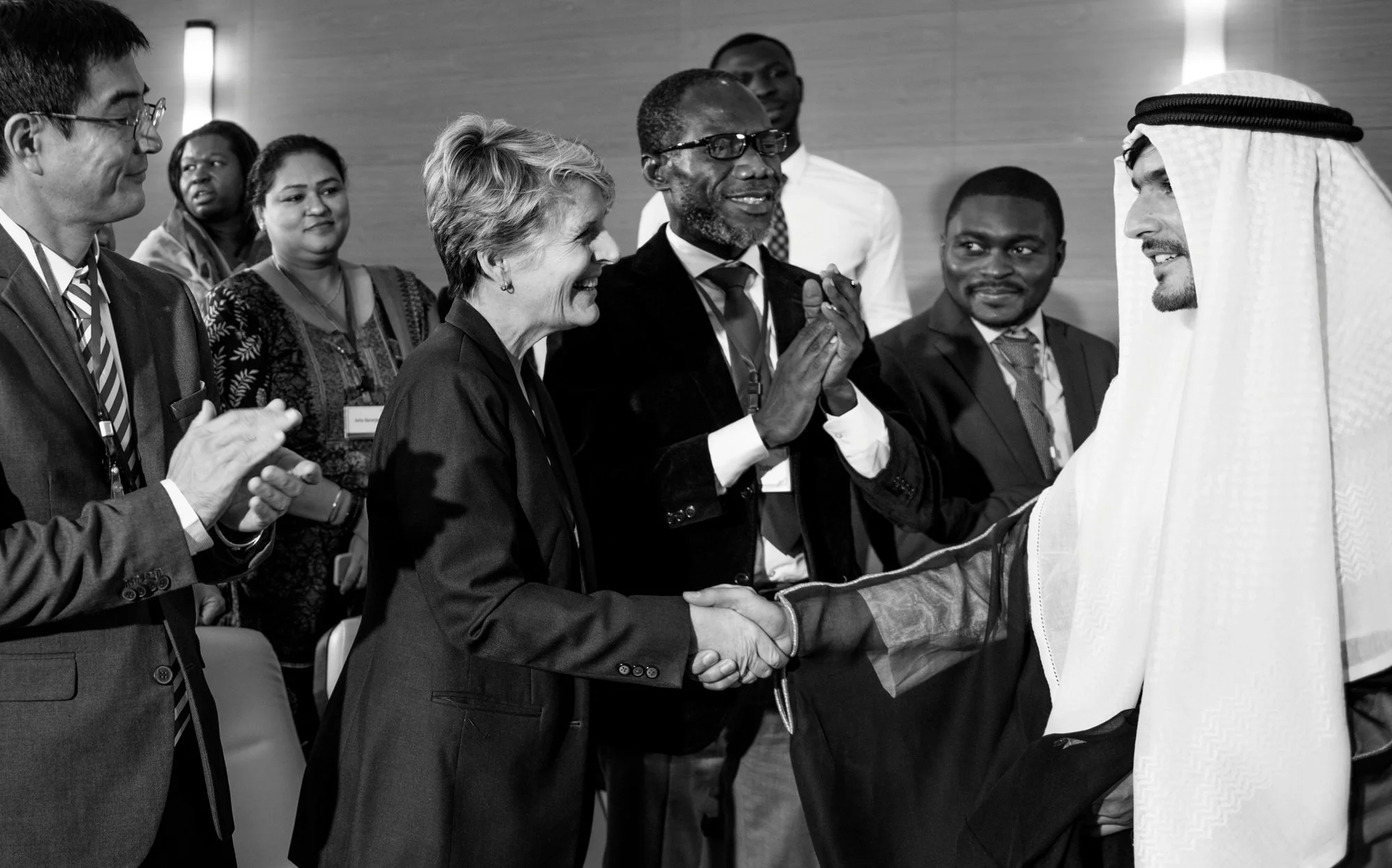 A group of diverse professionals, including a woman and man shaking hands, in a formal setting. The woman greets a man in traditional Middle Eastern attire, with others observing and smiling.