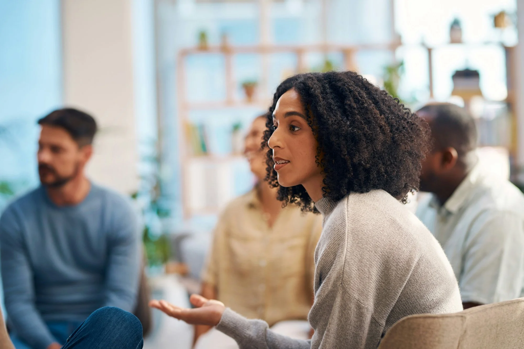 Group of diverse adults in a discussion, with a woman in the foreground speaking and gesturing with her hand, seated in a well-lit room with plants and shelves in the background.