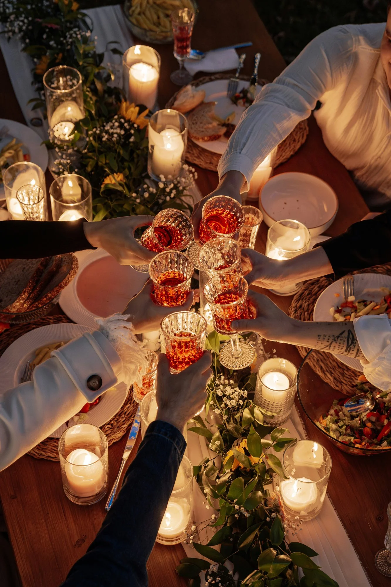 People raising glasses of rosé wine in a toast at a dinner party with candles and floral decorations on the table.
