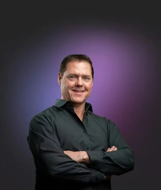 A man with short brown hair, smiling and crossing his arms, wearing a dark button-up shirt against a purple gradient background.