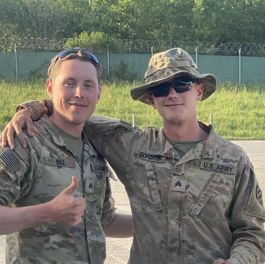 Two young men in U.S. Army combat uniforms standing outdoors, smiling, with one giving a thumbs-up and the other wearing sunglasses and a camouflage hat.