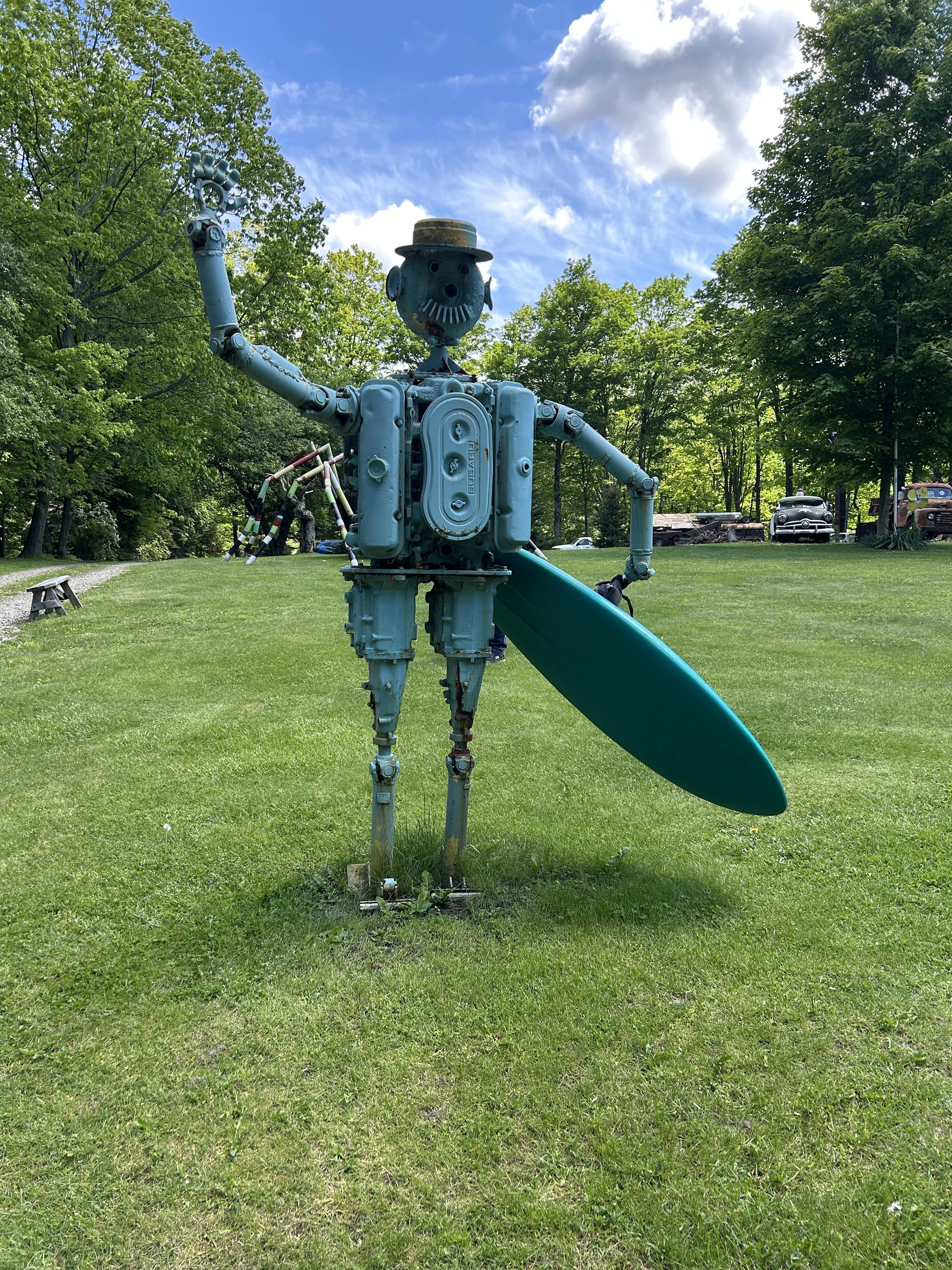A sculpture of a humanoid figure made from blue metal parts, holding a surfboard and wearing a small hat, standing on a grassy area with trees and parked cars in the background.