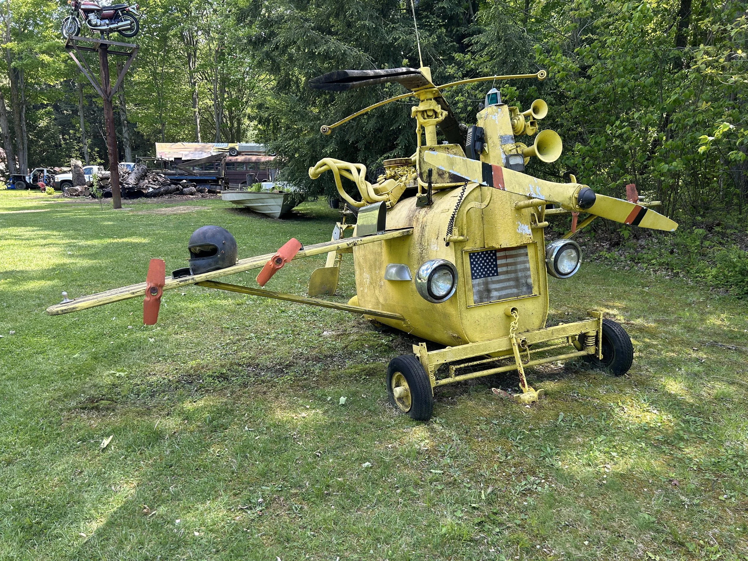 A yellow, custom-made helicopter with various unconventional modifications, parked on a grassy area with trees in the background.