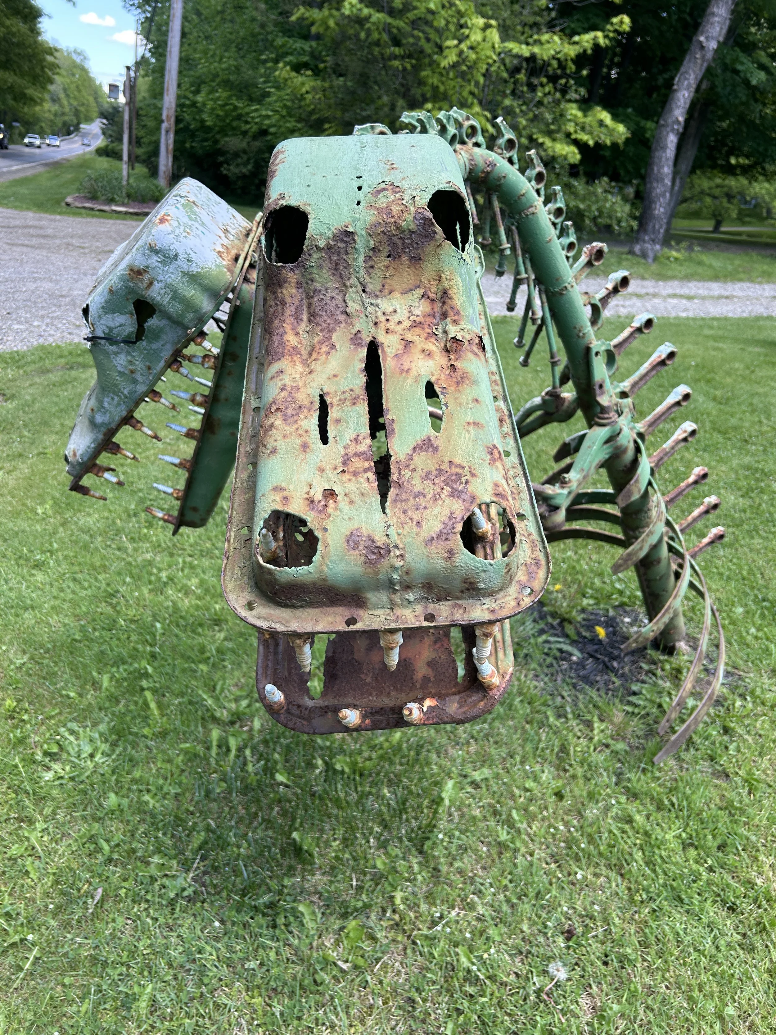 Rusty, abandoned mechanical sculpture resembling a creature with a skull face, made from old metal parts, situated outdoors on grass.