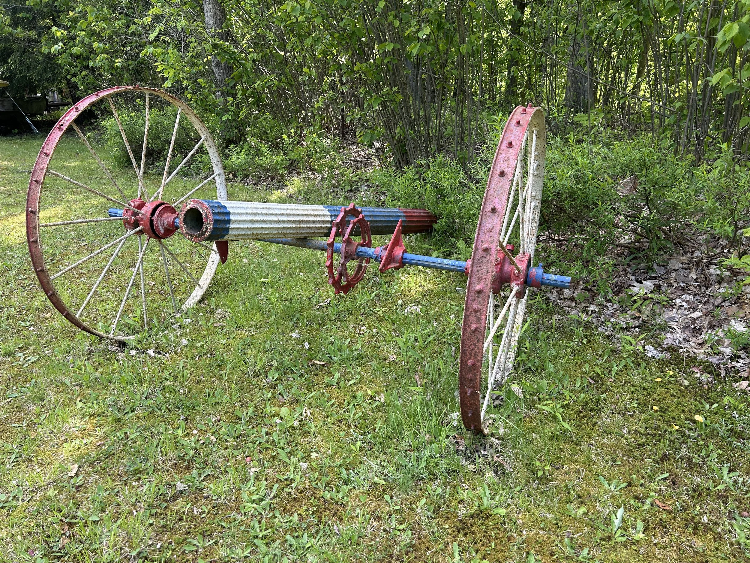 Rusty, vintage, red, and white farm wheelbarrow frame with blue accents, lying on grass in a wooded area.