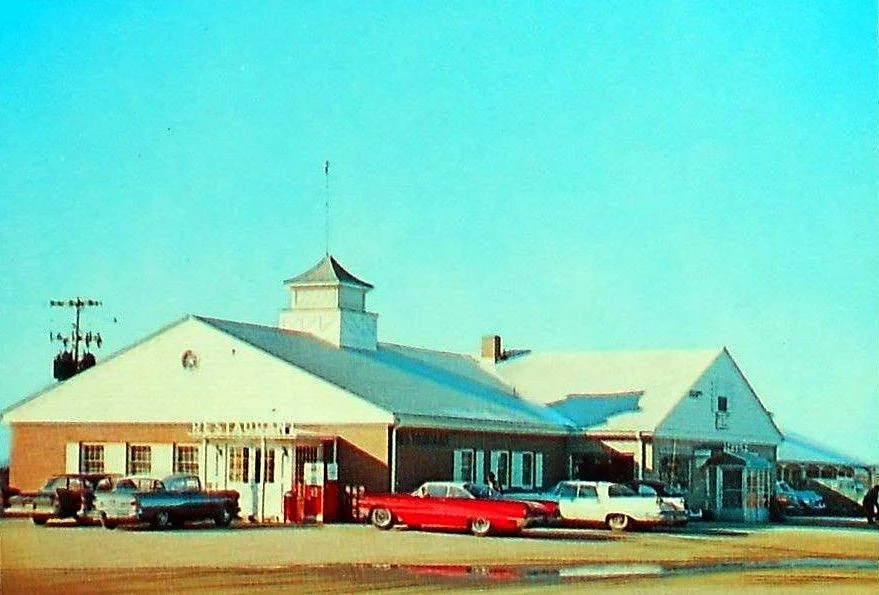 A vintage-style building, possibly a train station or town hall, with multiple cars parked in front, a clear blue sky, and a prominent cupola on the roof.
