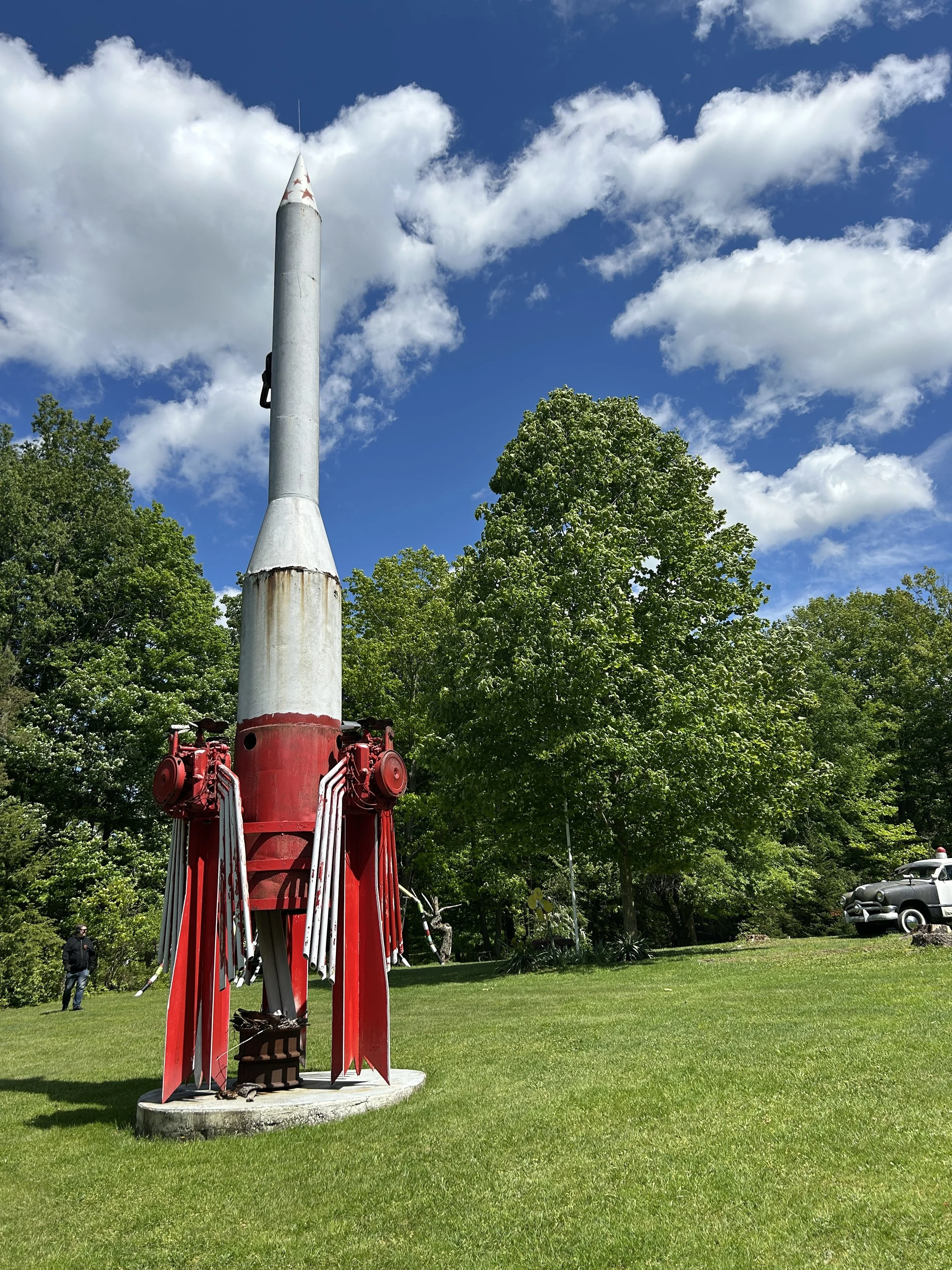 A vintage red and gray rocket statue on a grassy lawn with trees and a vintage car in the background, under a bright blue sky with scattered white clouds.