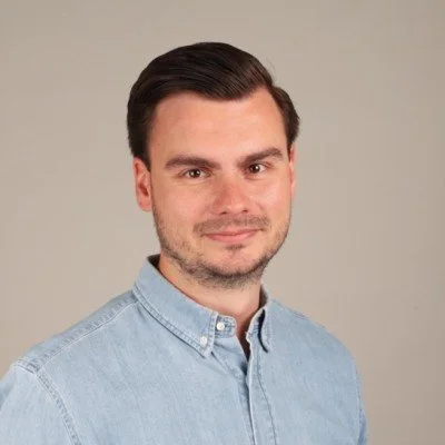 A man with dark brown hair and a light beard wearing a light blue button-up shirt, standing against a neutral background.
