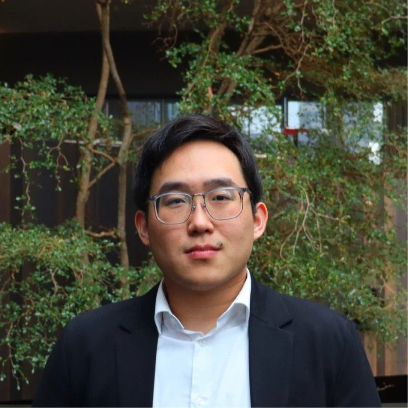 A young man wearing glasses and a formal black suit with a white shirt, standing outdoors in front of green trees and plants.