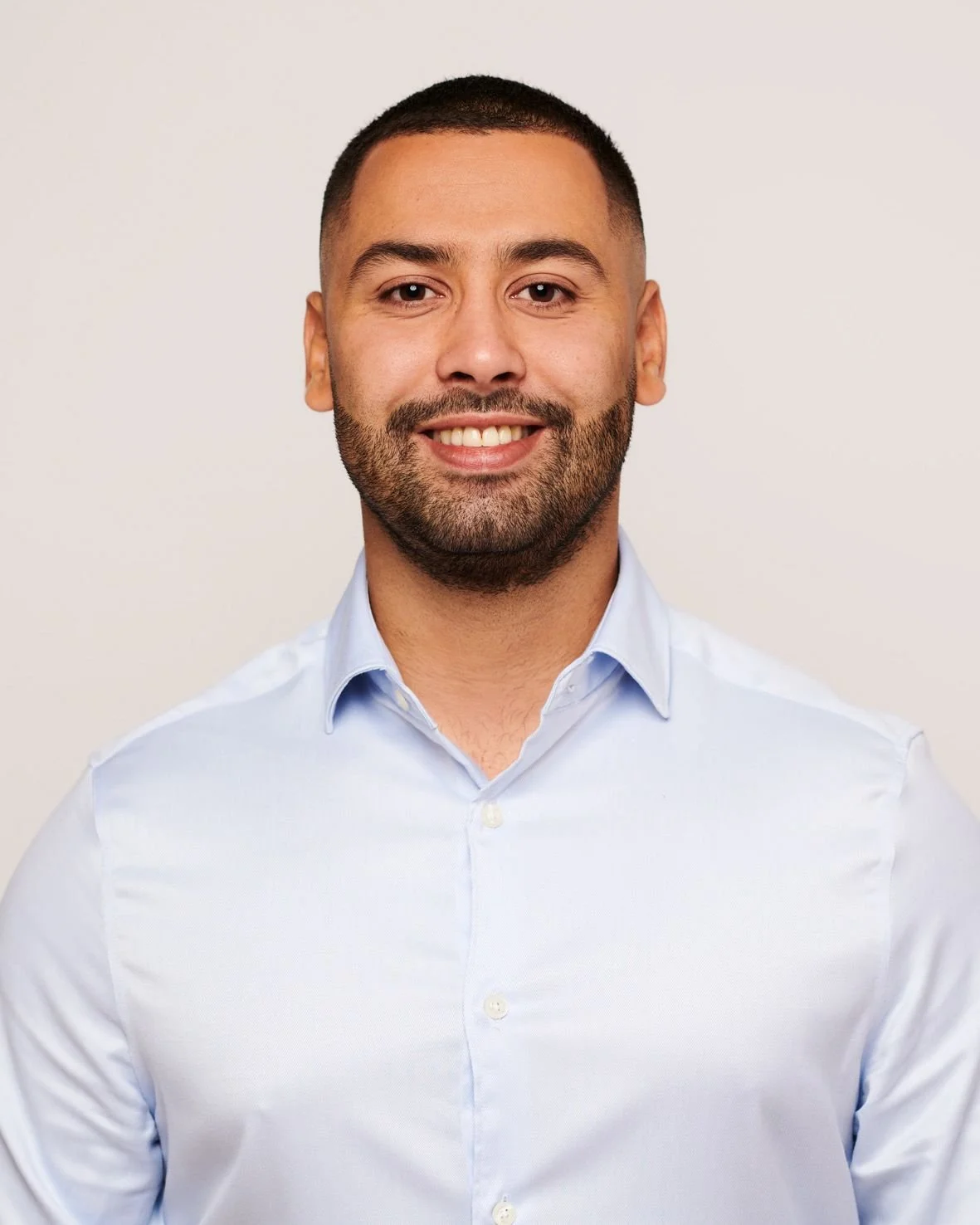 A smiling man with short dark hair and a beard, wearing a light blue dress shirt, standing against a plain light background.
