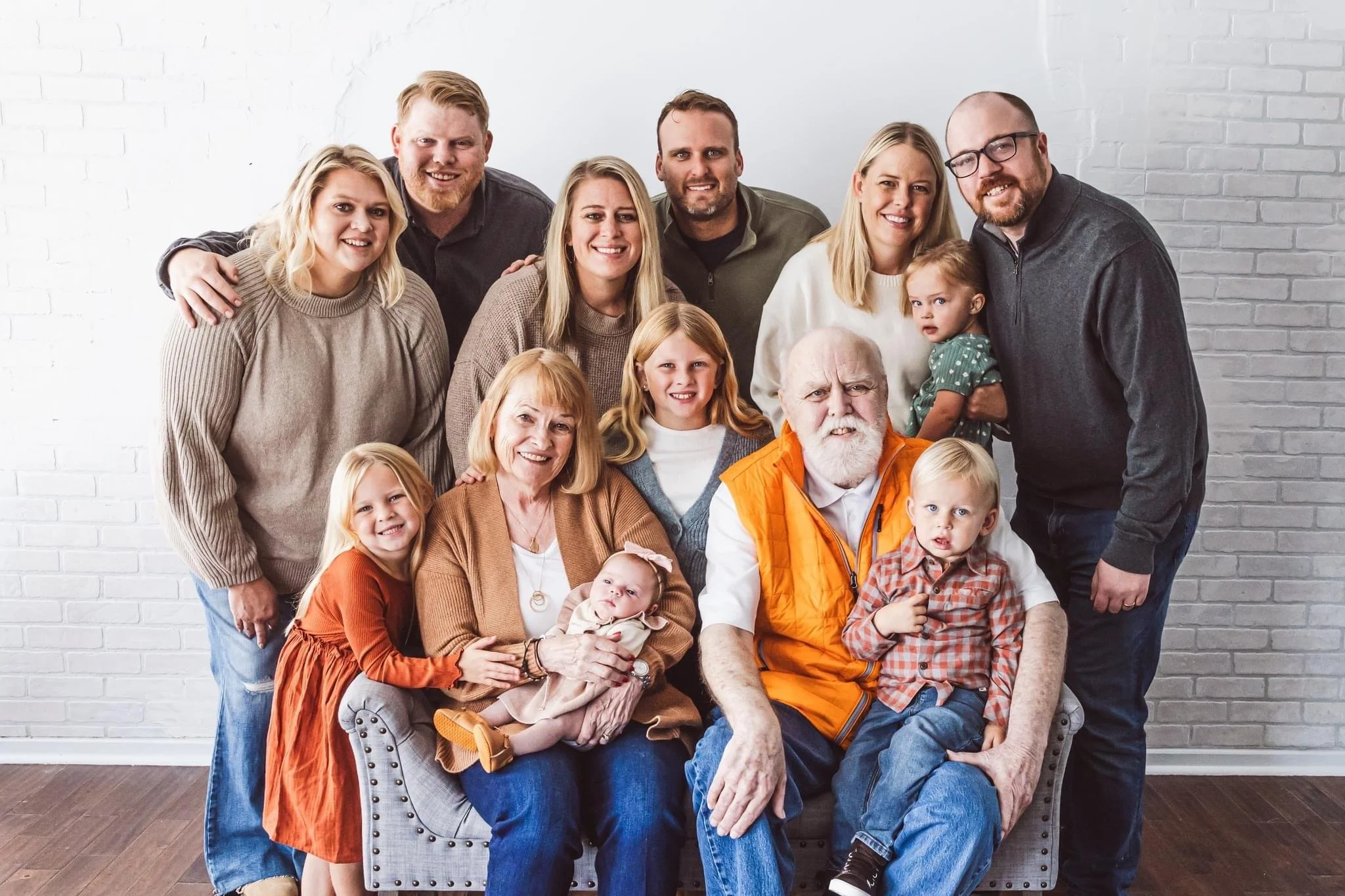 Family photo of multiple generations including grandparents, parents, and children posing in a room with a white brick wall background.