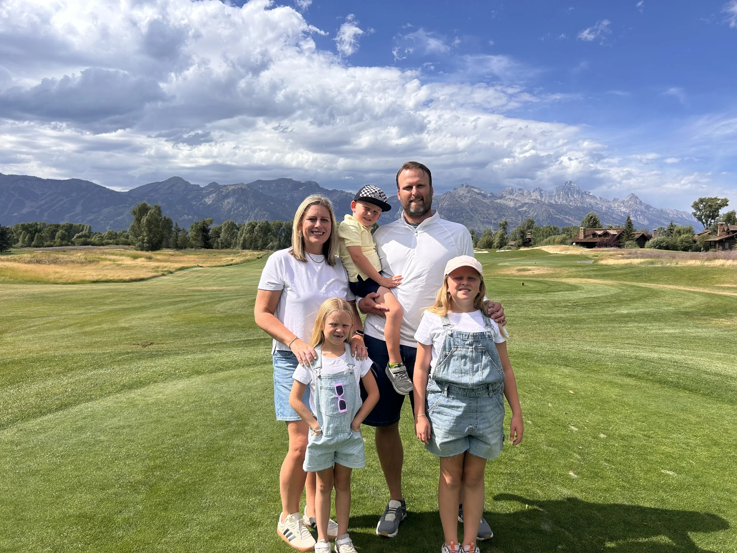A family of six, including two adults and four children, stands on a golf course with mountains in the background. The family is smiling and posing for the photo in a scenic outdoor setting.