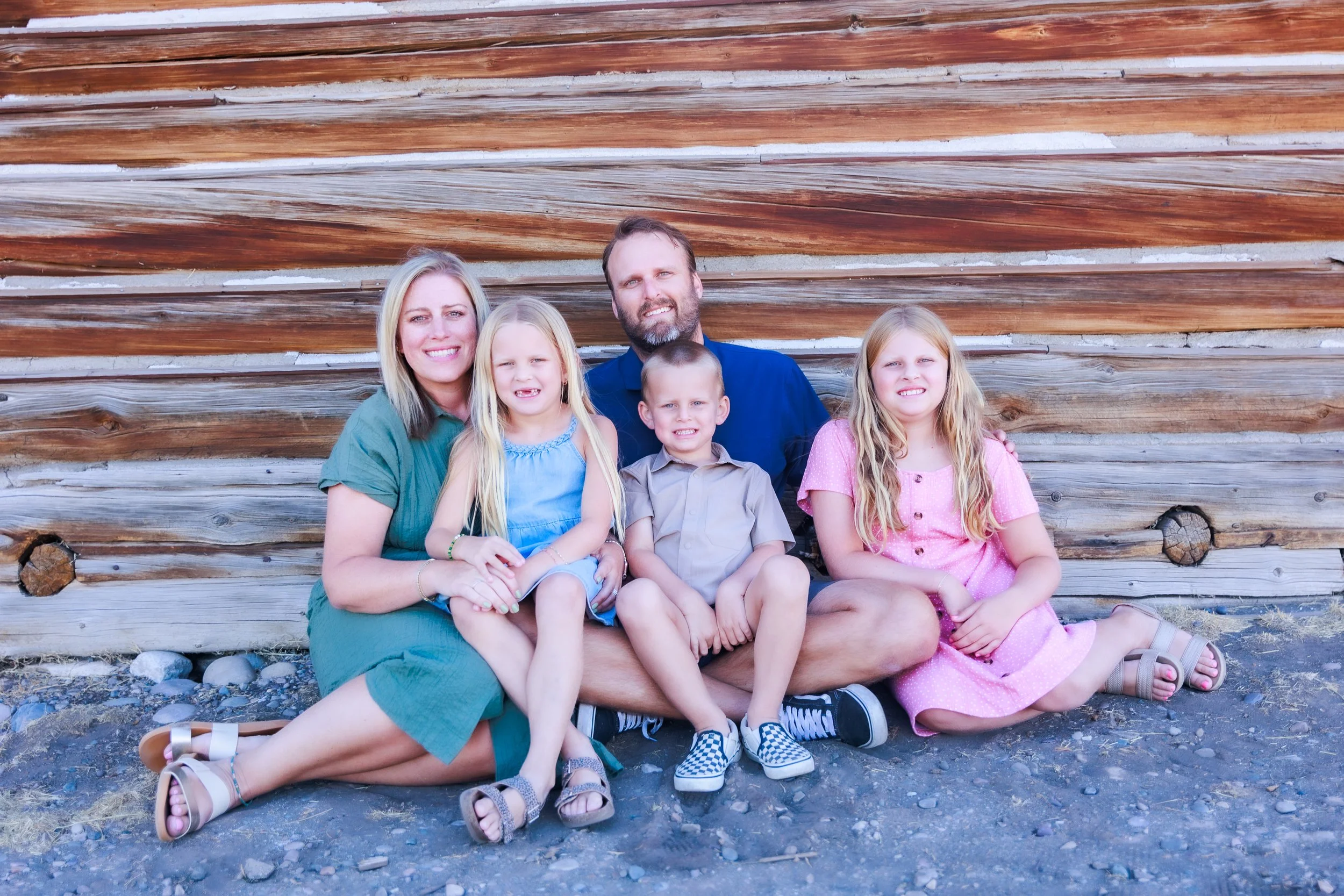 A family of five sitting on the ground in front of a rustic wooden wall, smiling at the camera. The group includes a woman, a man, two young girls, and a young boy.