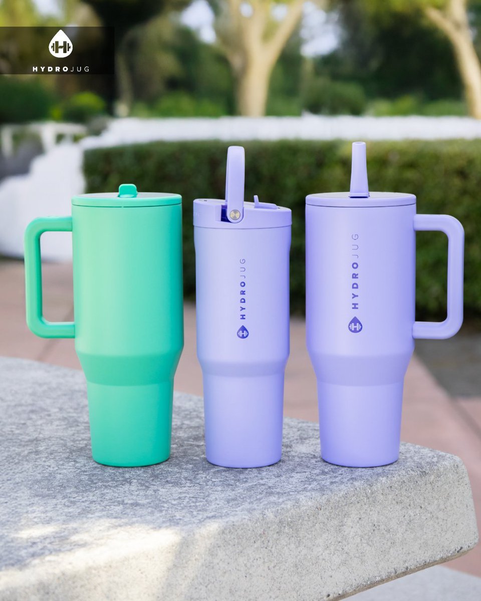 Three colorful reusable insulated travel mugs with straws, placed on a concrete surface outdoors with trees and bushes in the background. One mug is mint green, and two are lavender. The mugs feature the HydroJug logo.