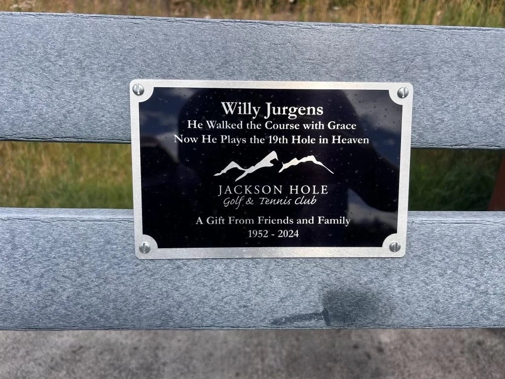 Plaque on park bench honoring Willy Jurgens, mentioning his walking the course with grace and now playing the 19th hole in heaven, indicating he was associated with Jackson Hole Golf & Tennis Club, and noting it was a gift from friends and family from 1952 to 2024.
