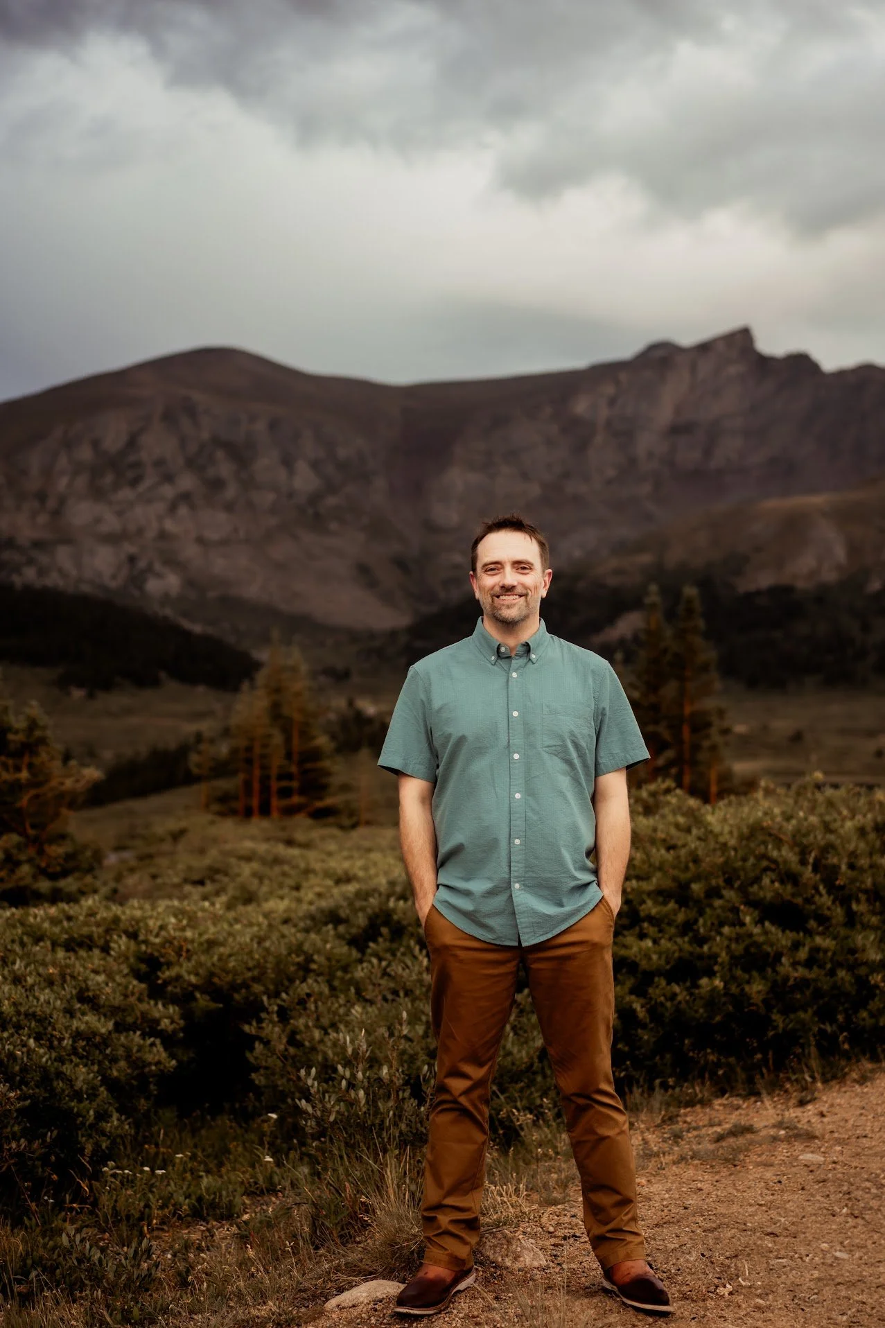 A smiling man with short brown hair and stubble, wearing a teal button-up shirt and brown pants, stands outdoors on a dirt trail in front of mountains and trees under a cloudy sky.
