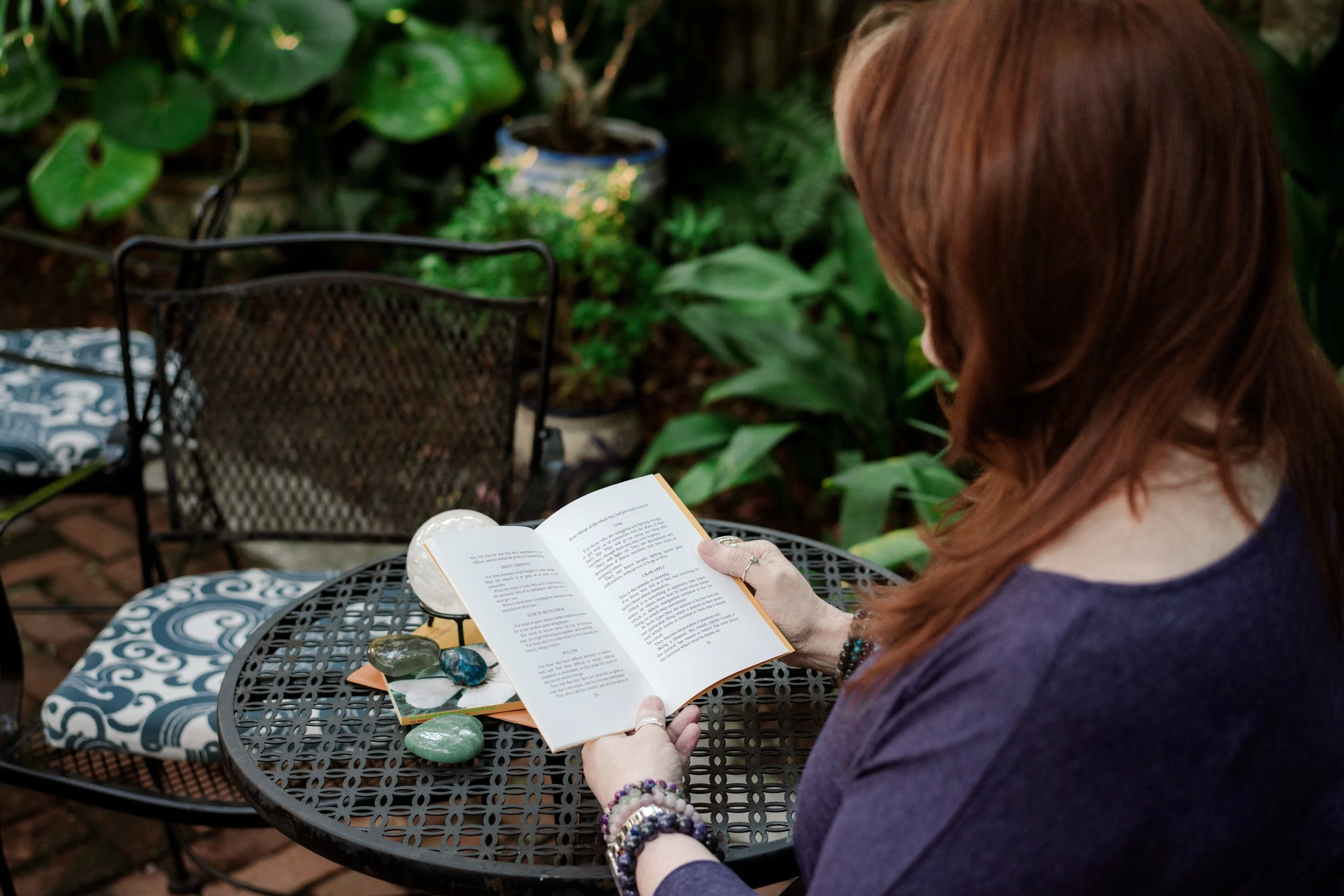 Woman with red hair reading a book at an outdoor table decorated with crystals and gemstones in a garden setting.