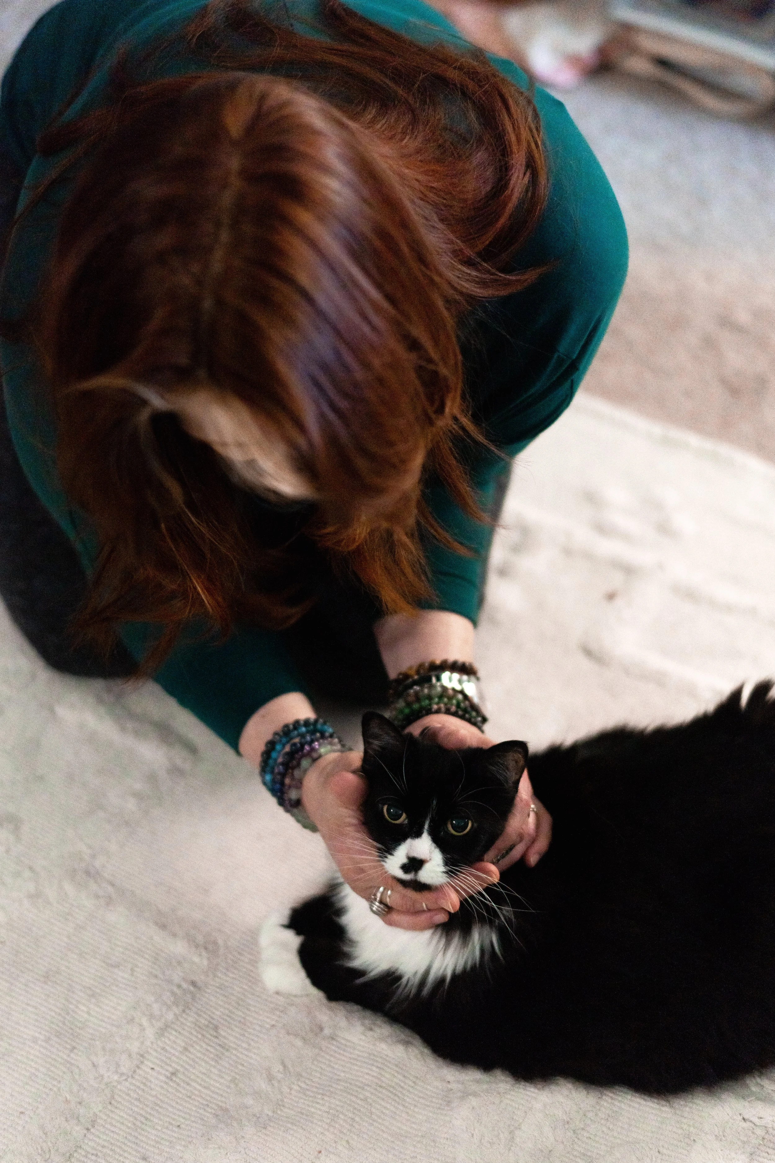 A woman with reddish-brown hair, wearing bracelets, gently holding a black and white cat while crouching on a light-colored carpet.