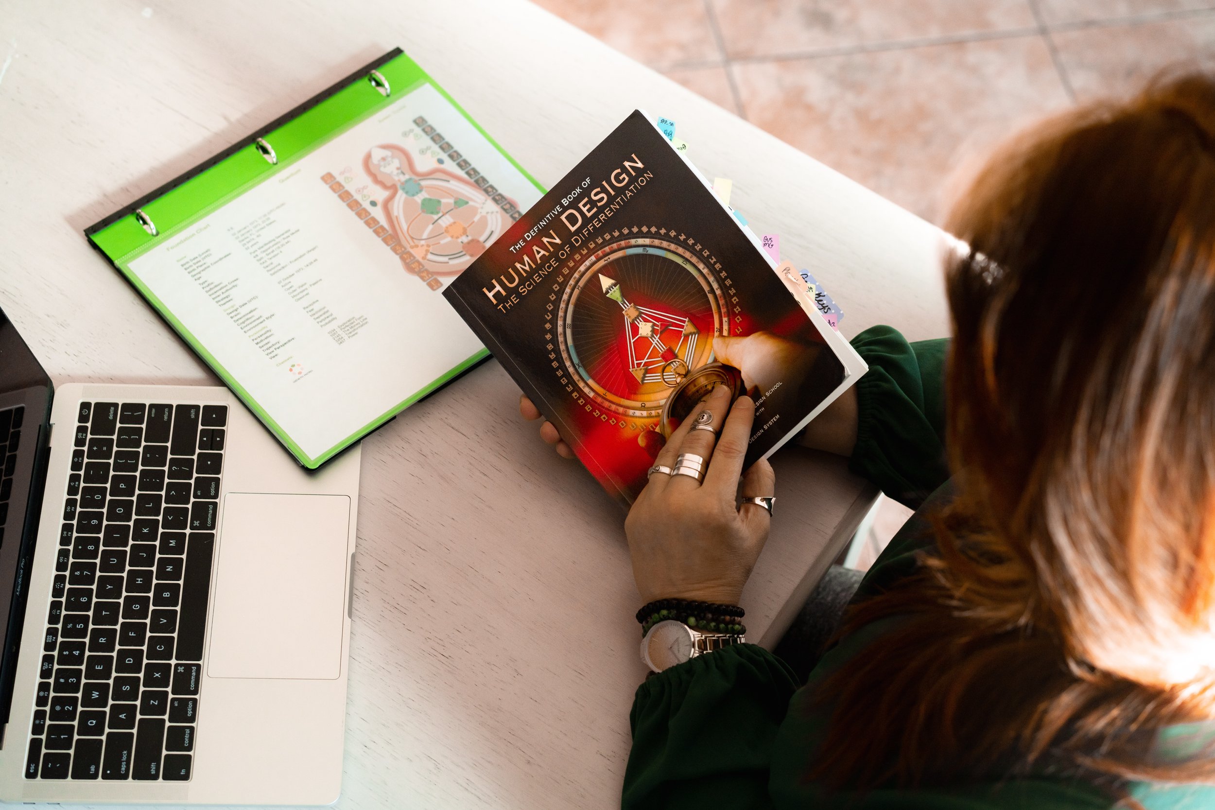Person with reddish-brown hair reading a book titled 'The Definitive Book of Human Design' at a white desk with an open laptop and a green binder.