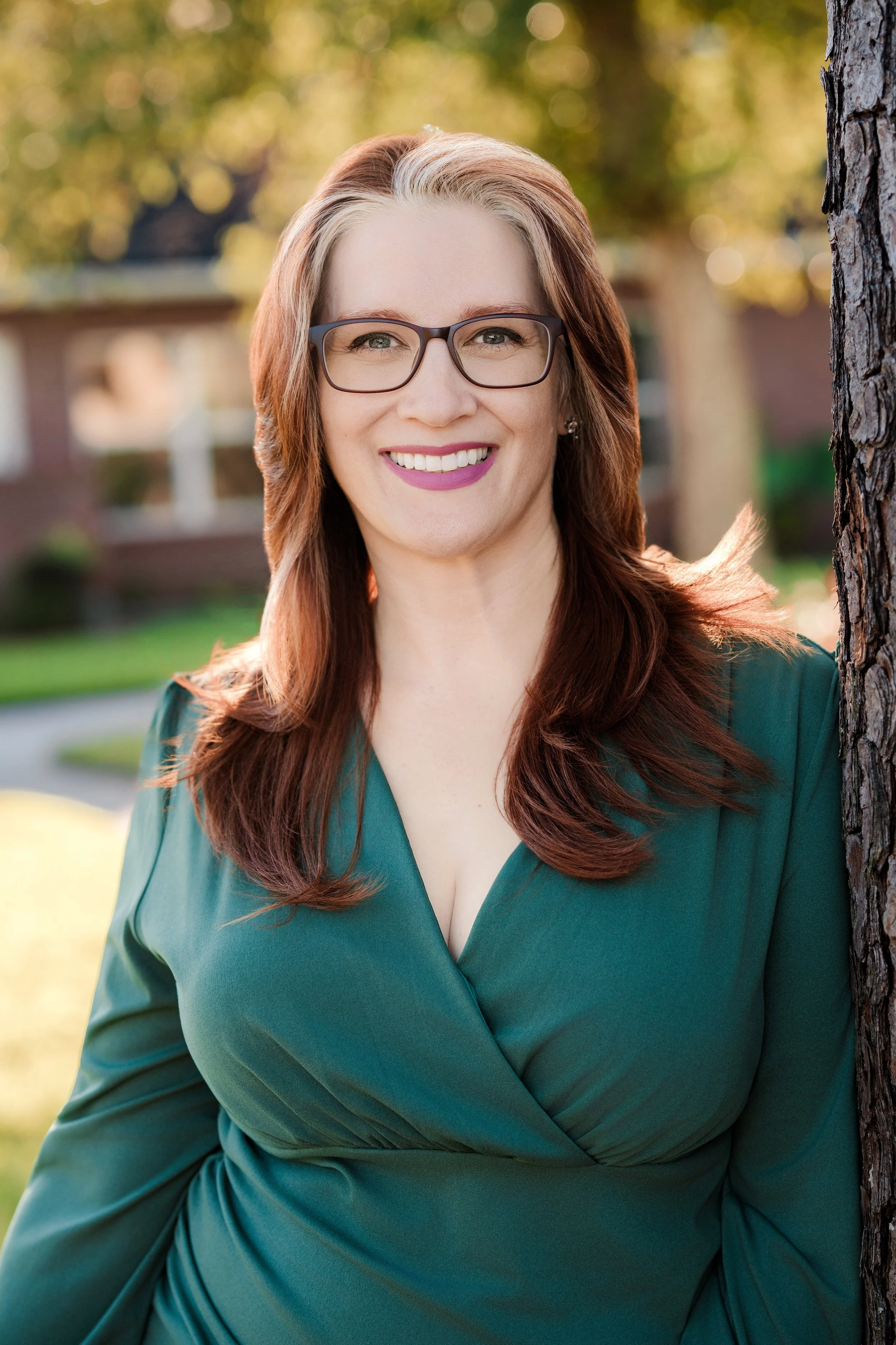 A woman with red hair, glasses, and a green dress smiling outdoors next to a tree with a blurred park background.