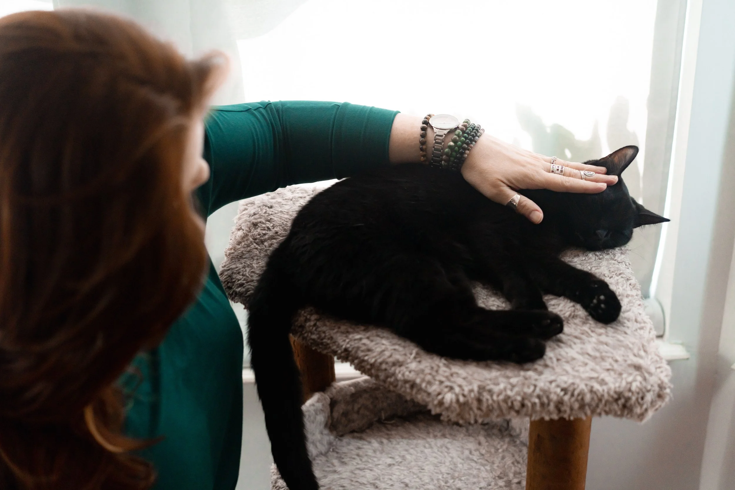 A woman petting a black cat lying on a soft, beige scratching post platform near a window.