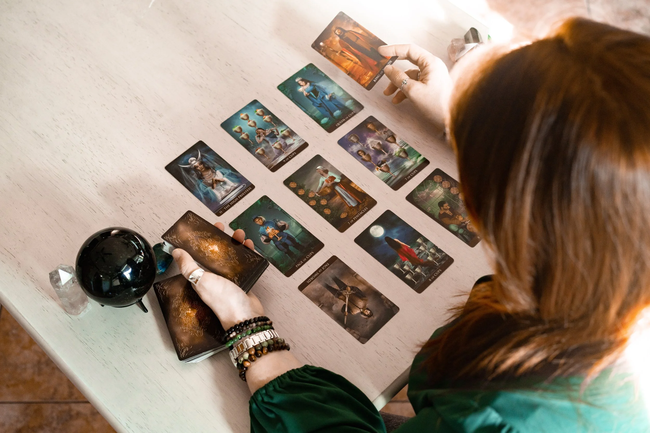 A person with reddish-brown hair and wearing a green long-sleeve shirt is sitting at a table, spreading out tarot cards, with a black sphere and a small crystal on the table. The person has multiple bracelets on their left wrist.