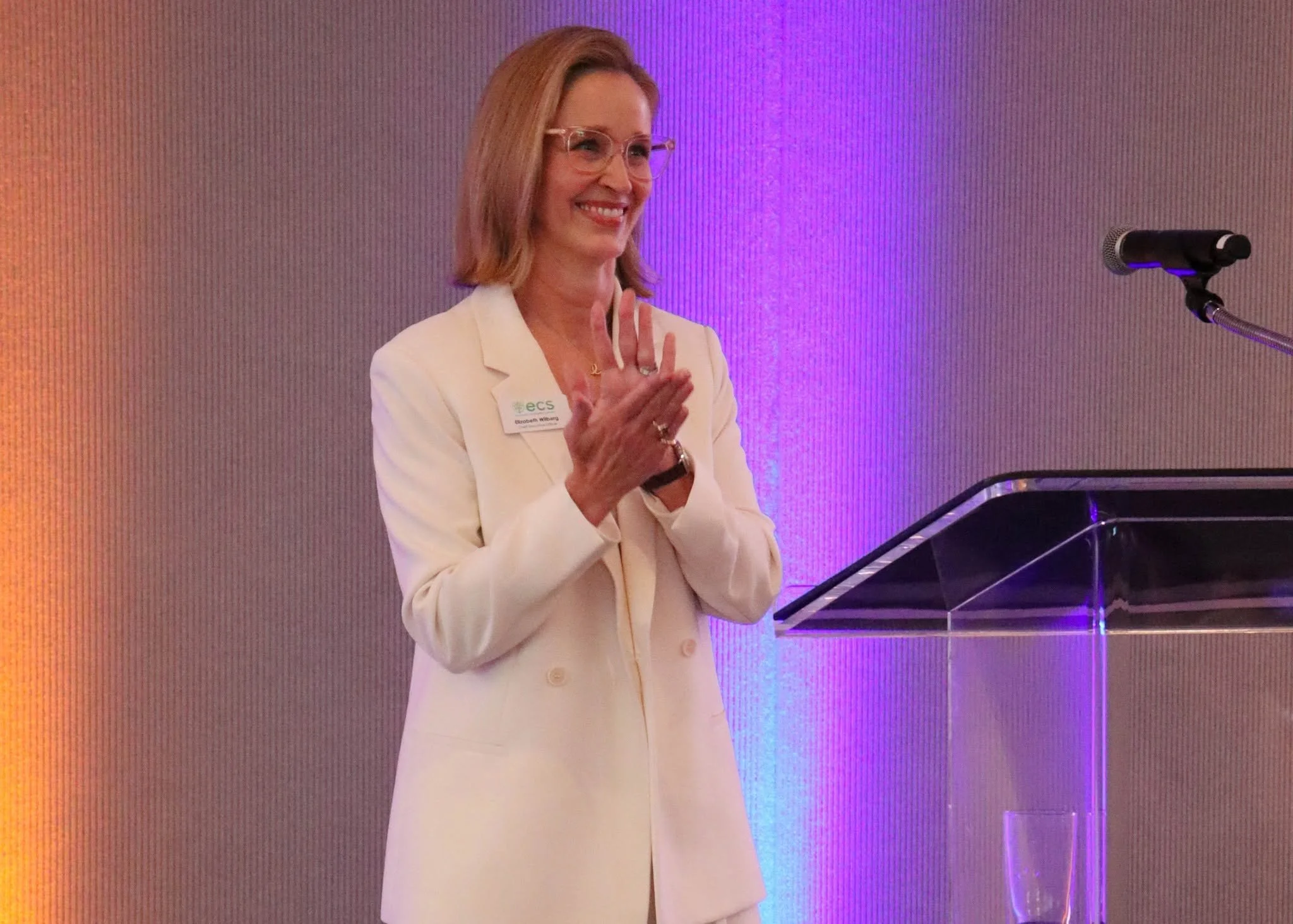 A woman with blonde hair and glasses, wearing a white blazer with a name tag, standing at a podium with a microphone in front of a purple-lit background, making a hand gesture.
