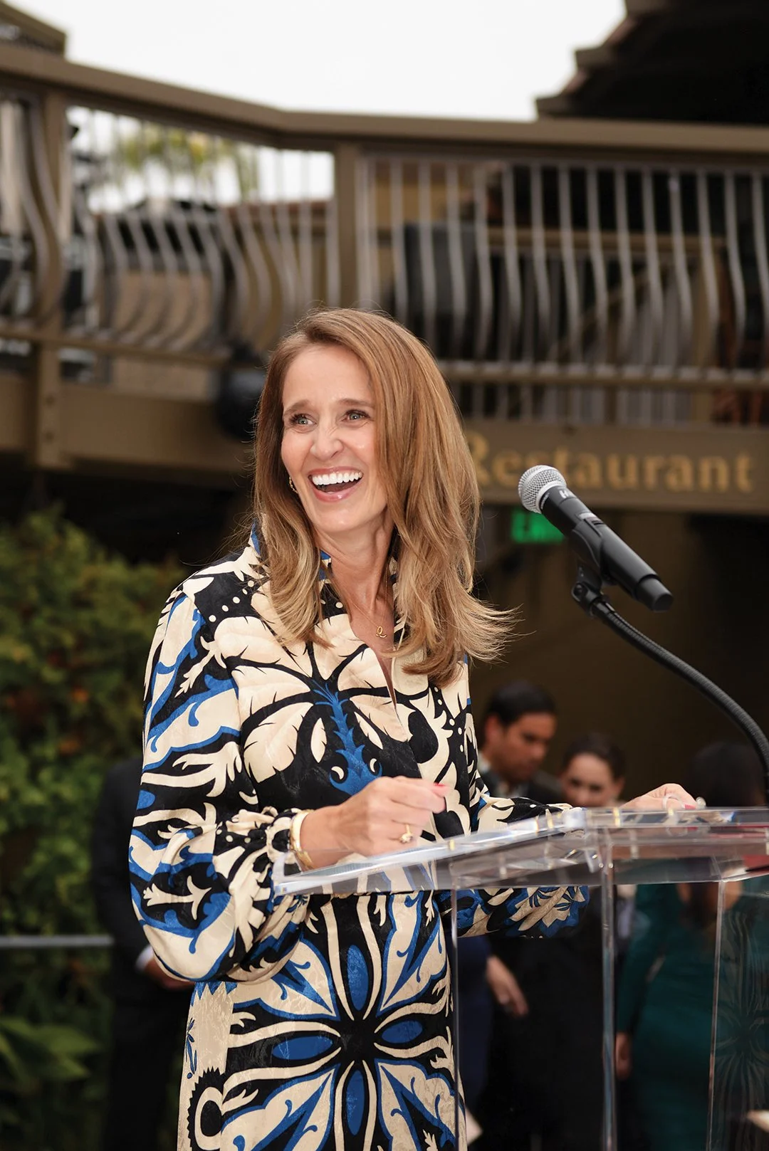 A woman with light brown hair smiling and speaking at a microphone during an outdoor event, wearing a black, white, and blue patterned dress.
