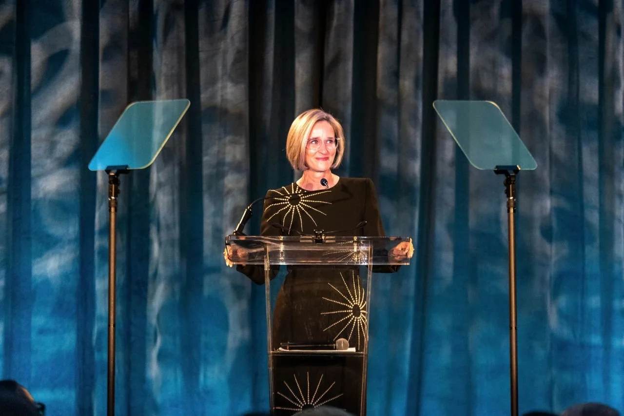 A woman with blonde hair standing behind a clear podium on a stage, with a blue curtain backdrop and two teleprompters on either side.