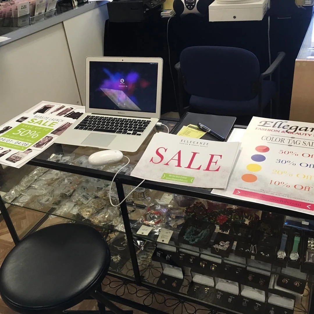 A table with sale signs, a laptop, a mouse, a notepad, and a pen in a retail store. The glass display case below contains jewelry and watches. There are chairs near the table.
