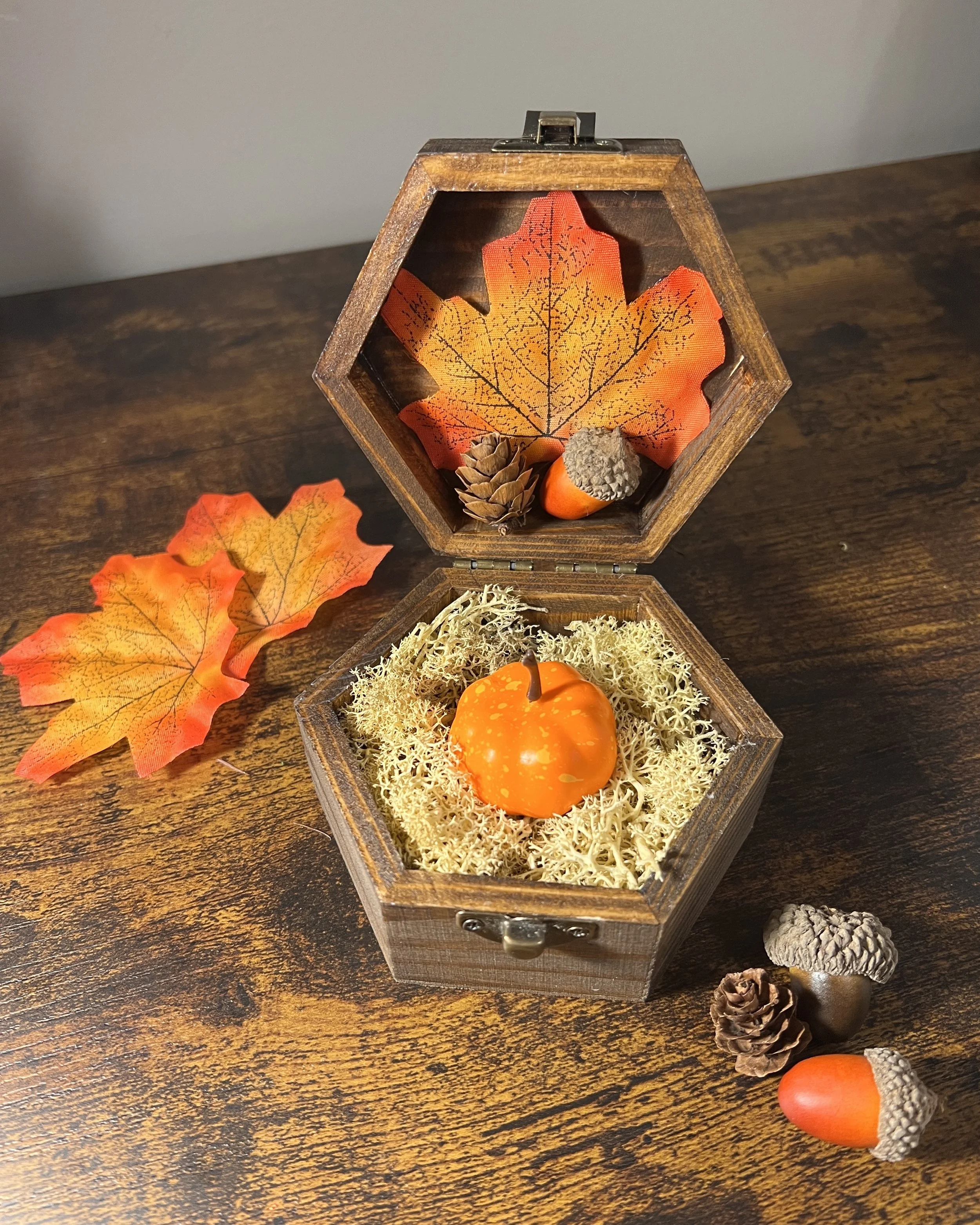 Wooden, brown hexagonal hinged box with autumn leaf, pinecone, and acorn displayed on the open lid and an orange pumpkin nestled in moss.