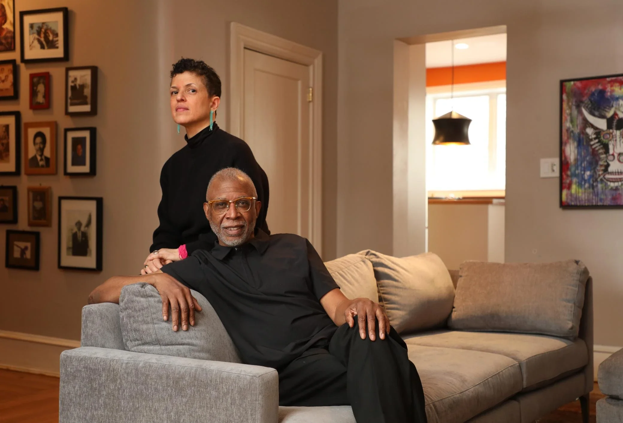 An older Black man with glasses and a gray beard sitting on a gray couch in a cozy living room, with a woman standing behind him. The woman has short curly hair and wears a black turtleneck. The wall behind them has framed photographs, and there is a colorful painting on the adjacent wall. A doorway reveals a room with a window and a black pendant light.