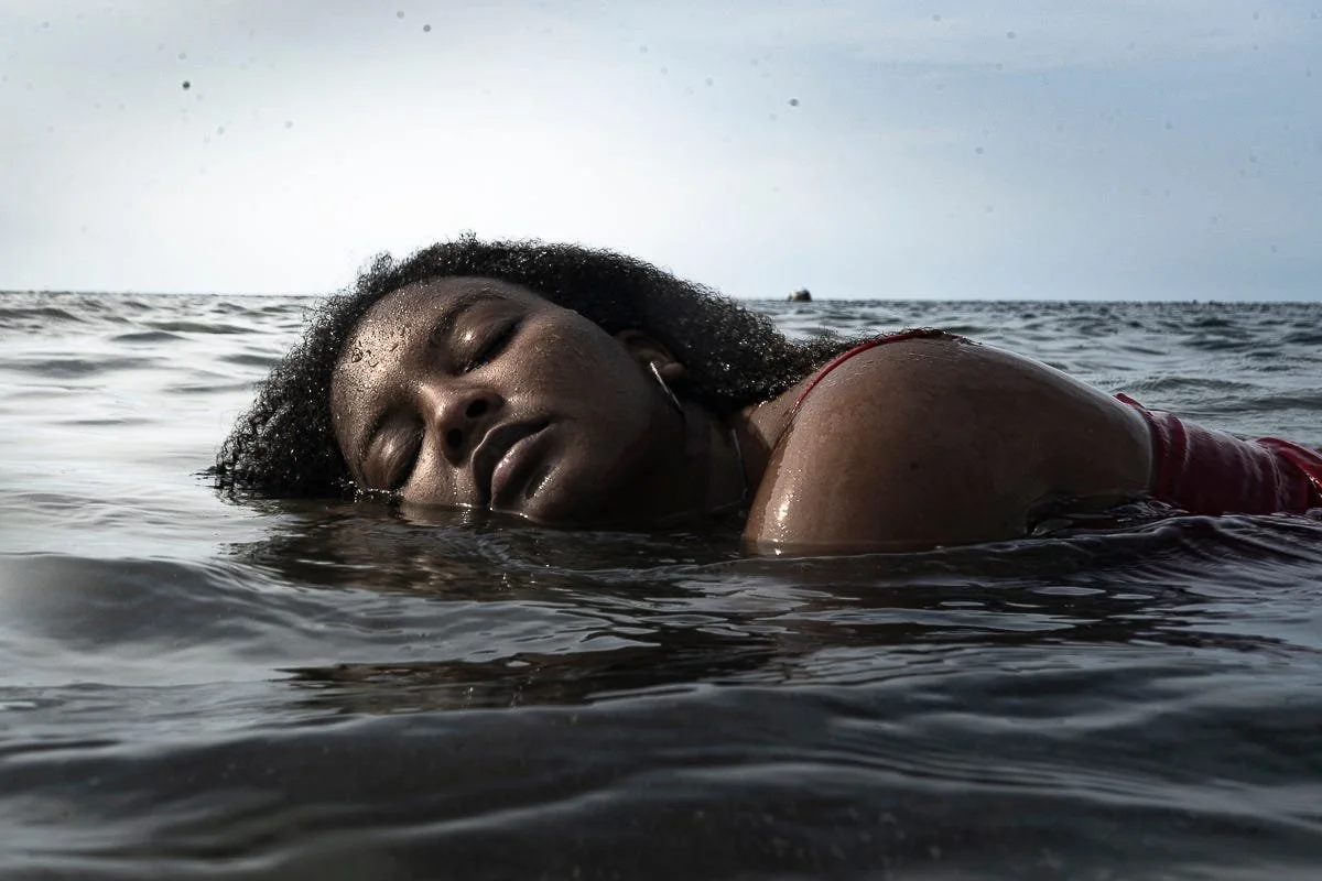 An AfroMexican woman with curly hair floating face down in the water at the ocean, with only her head and part of her body visible.