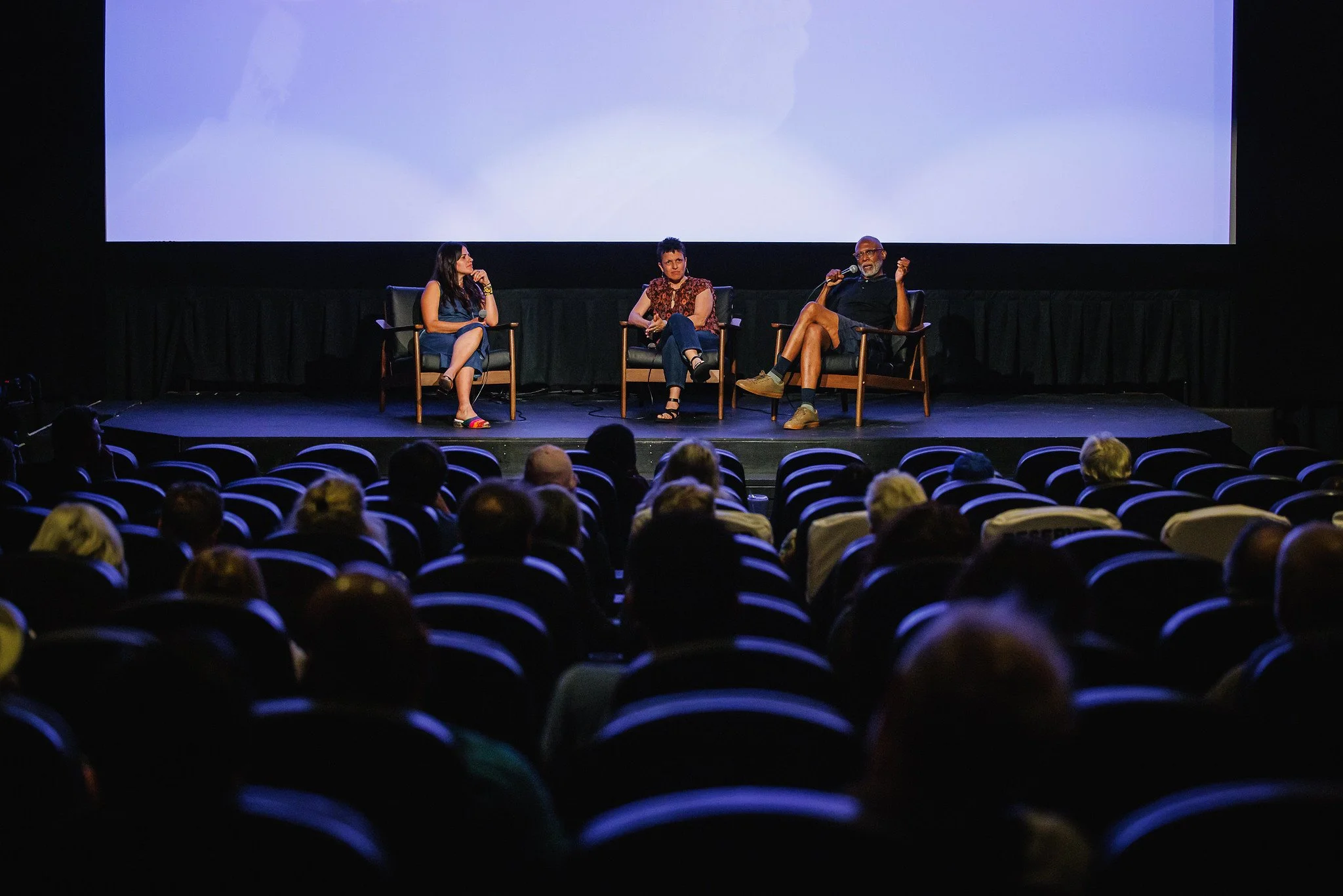 Three people are sitting on stage in a panel discussion, facing an audience, with a large screen behind them in a theater setting.