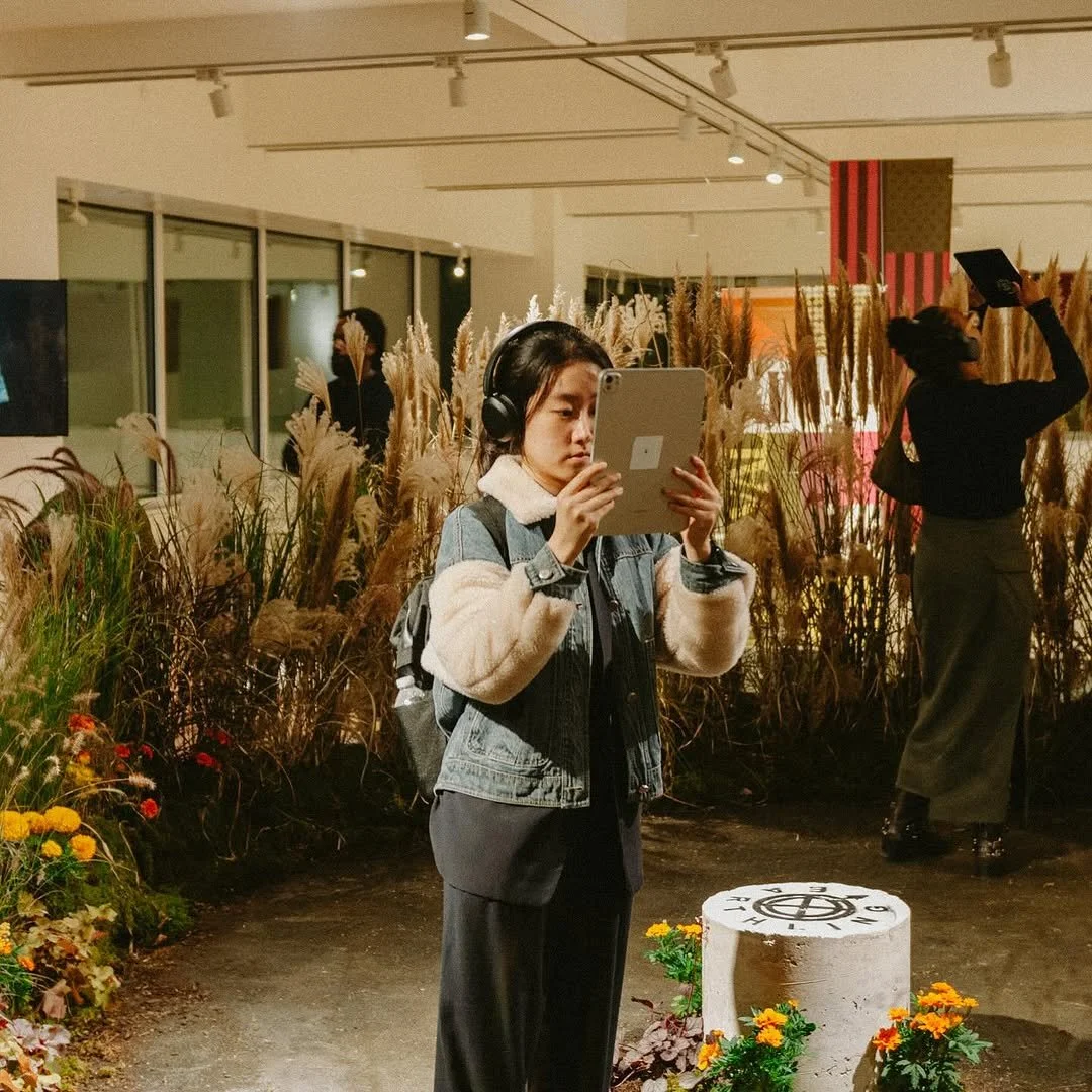A woman with headphones looking at her phone in an indoor area decorated with tall grasses and flowers. There are other people in the background, some standing and some with raised arms, in a setting with bright lighting and large windows.