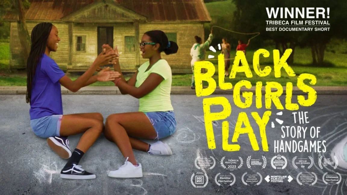 Two young black girls are playing hand games on an outdoor street, sitting on the ground and smiling at each other, with a house and trees in the background. The image has text indicating they won a film festival award for a documentary short called 'Black Girls Play: The Story of Handgames.'