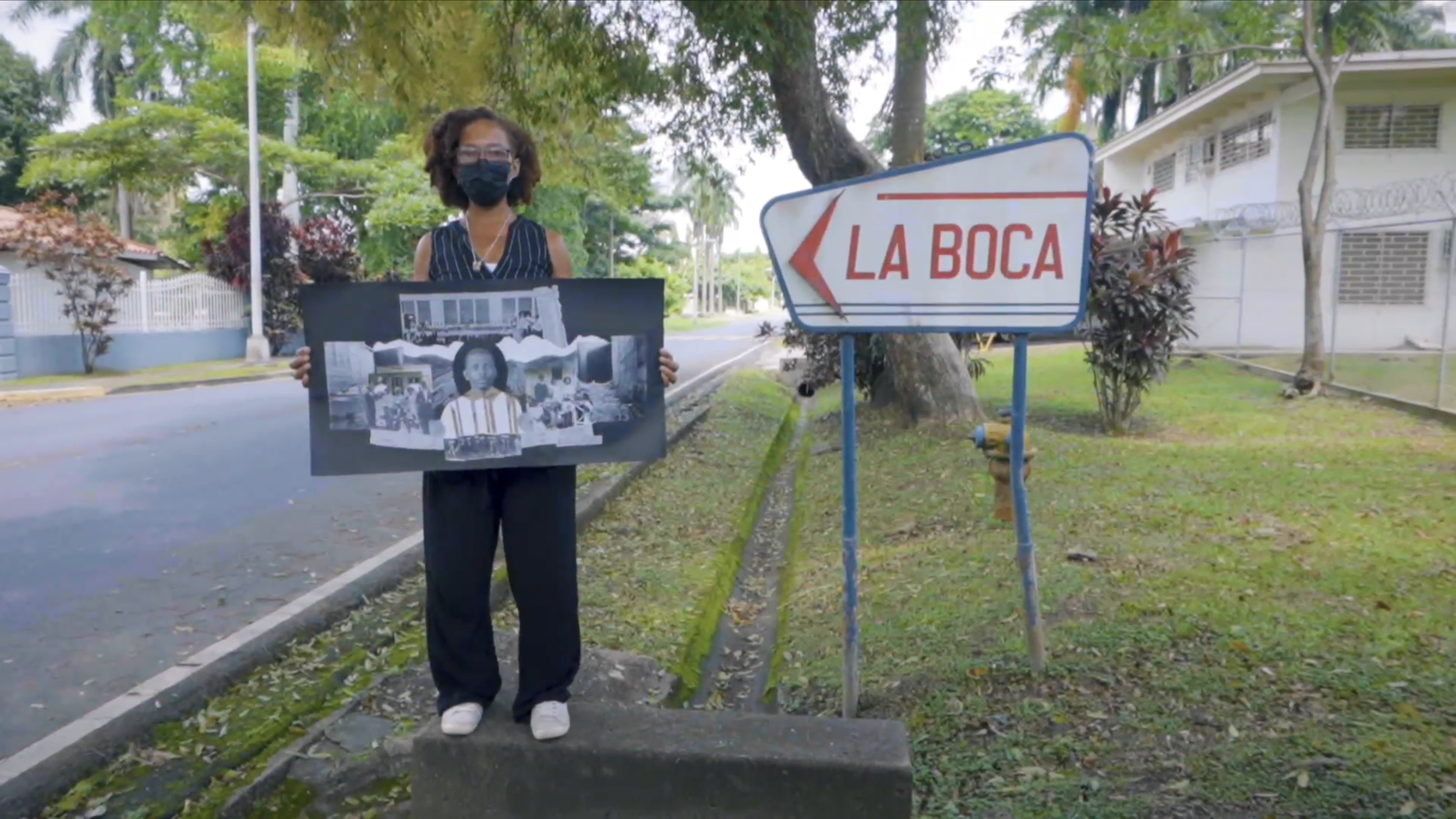 A woman wearing a black face mask holding a poster stands on a sidewalk beside a street with trees and houses in the background. There is a sign saying 'LA BOCA' with an arrow pointing left.