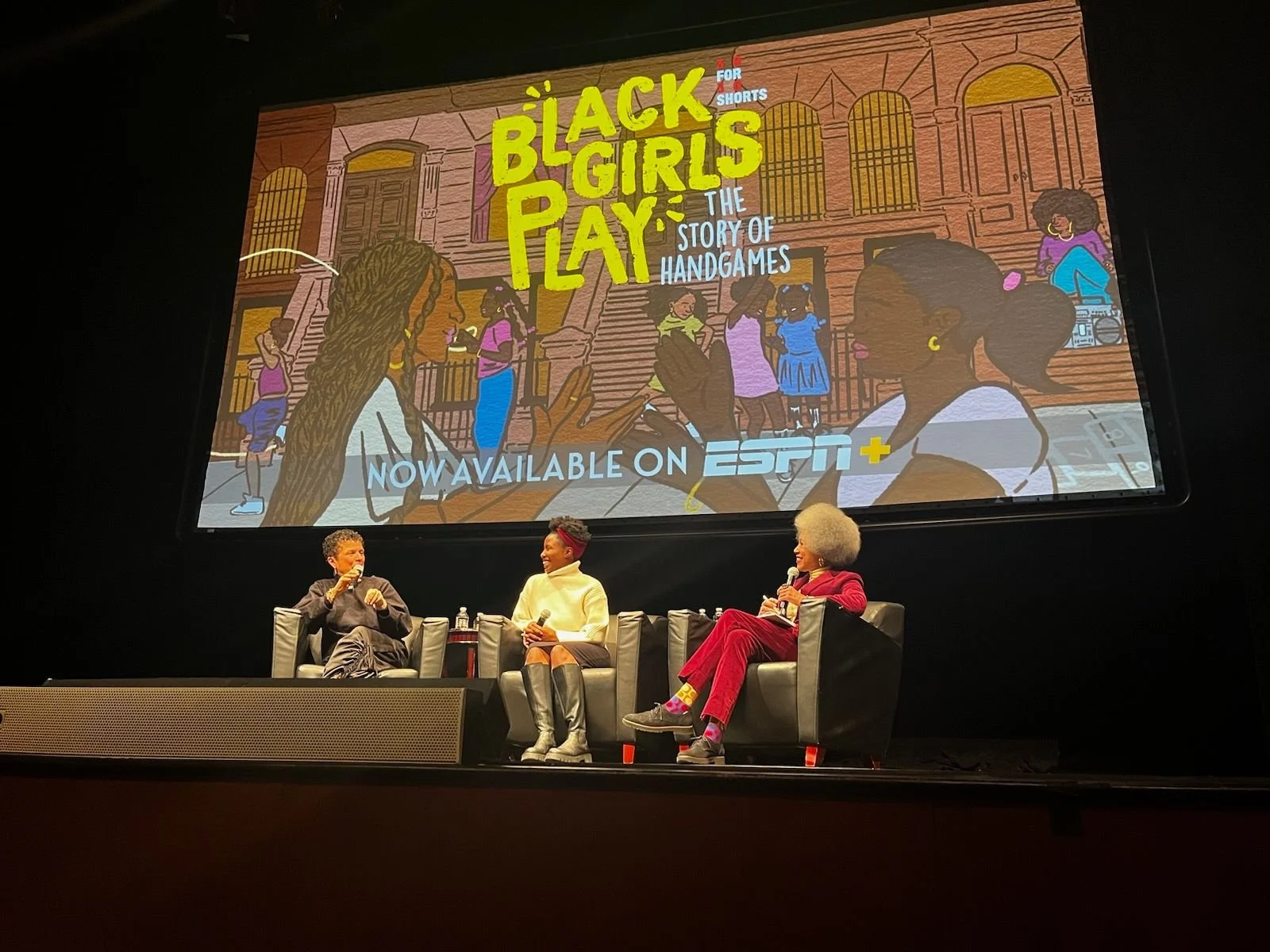Three women seated (among them director Michele Stephenson) on stage in front of a large screen, participating in a discussion or panel. The screen displays an animated poster for 'Black Girl Play: The Story of Handgames,' available on ESPN+.