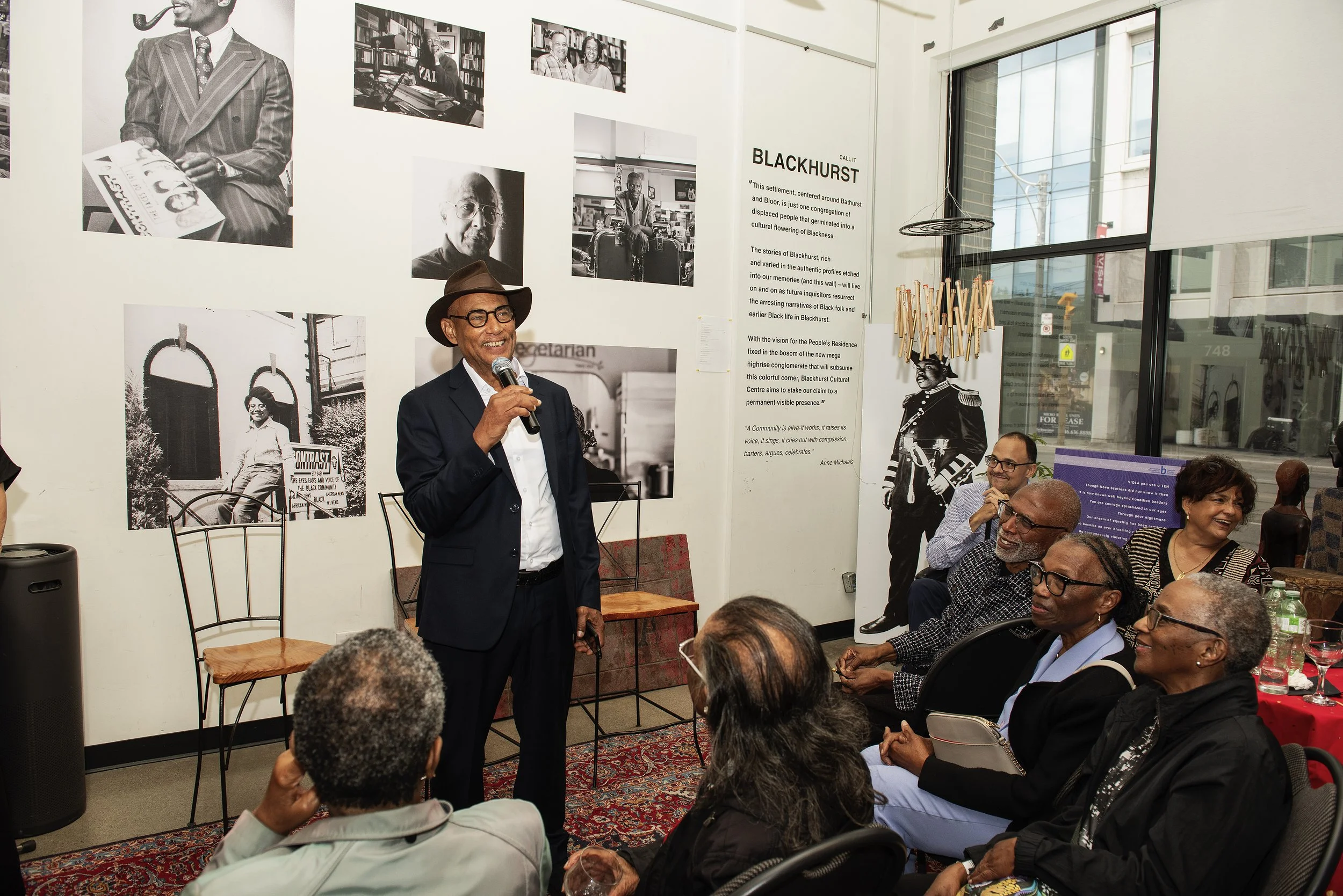 A man wearing a hat and glasses is speaking into a microphone at a gathering, with an audience of diverse adults watching him. The event is in a room with black-and-white photos and a large text panel on the wall, and a large window in the background.