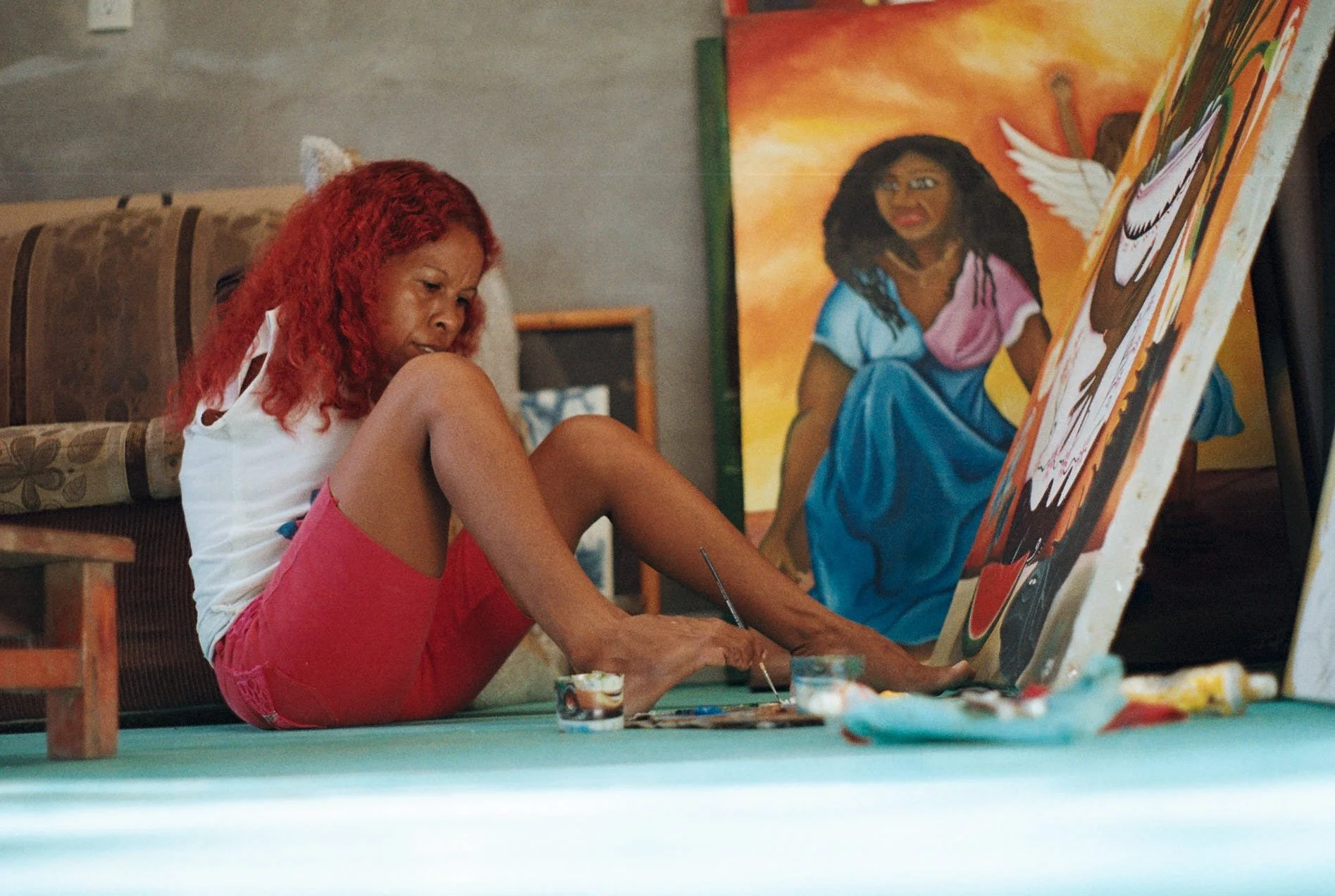 A AfroMexican woman with red hair, wearing a white tank top and pink shorts, sitting on the floor and painting a colorful portrait of a woman with dark curly hair, wearing a blue dress and pink shawl, against an orange background.
