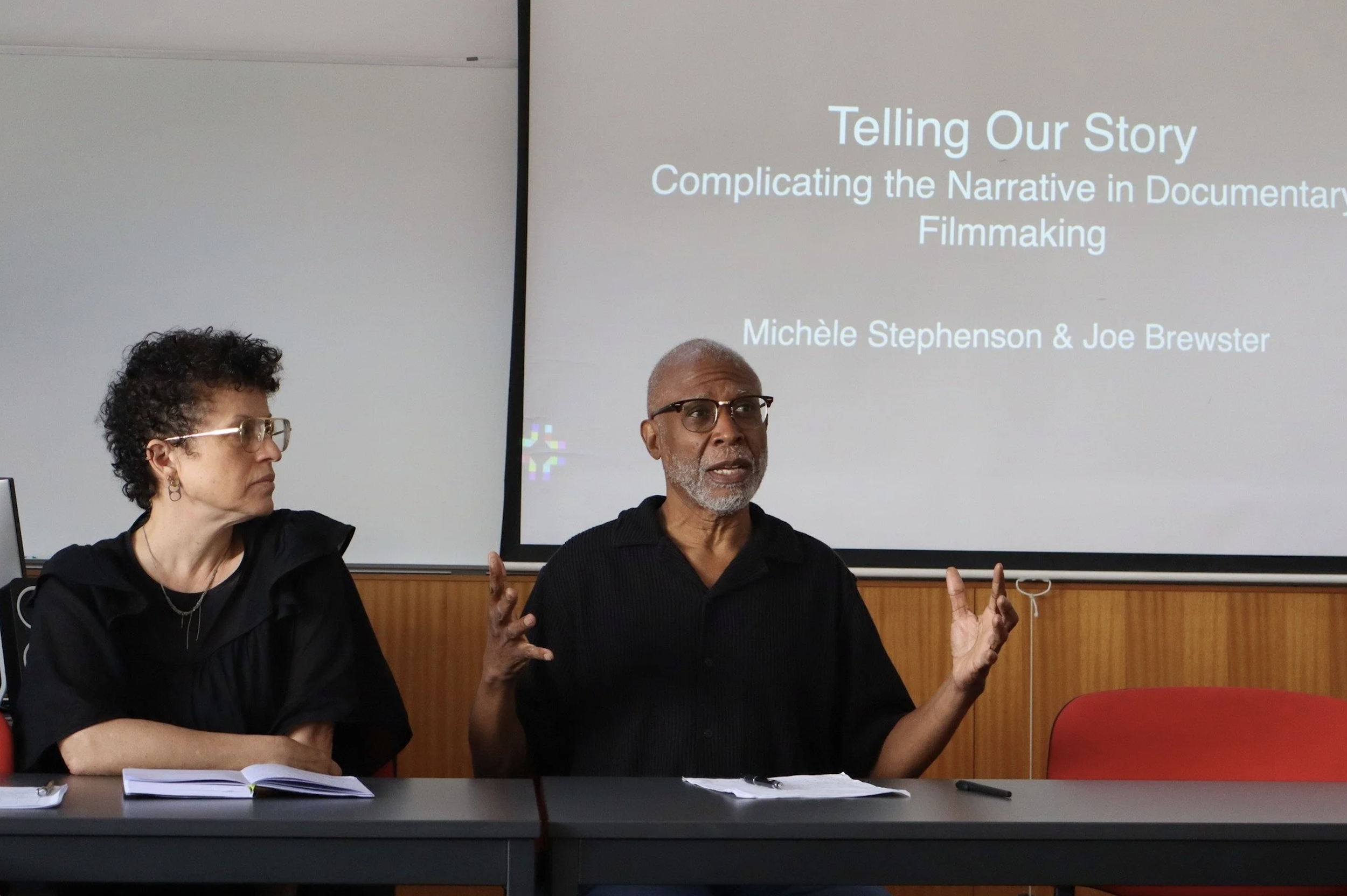 Two speakers, a woman on the left and a man on the right, seated at a table during a presentation. The woman is listening, while the man is speaking with hand gestures. A presentation slide behind them reads 'Telling Our Story: Complicating the Narrative in Documentary Filmmaking' with the names 'Michèle Stephenson & Joe Brewster'.