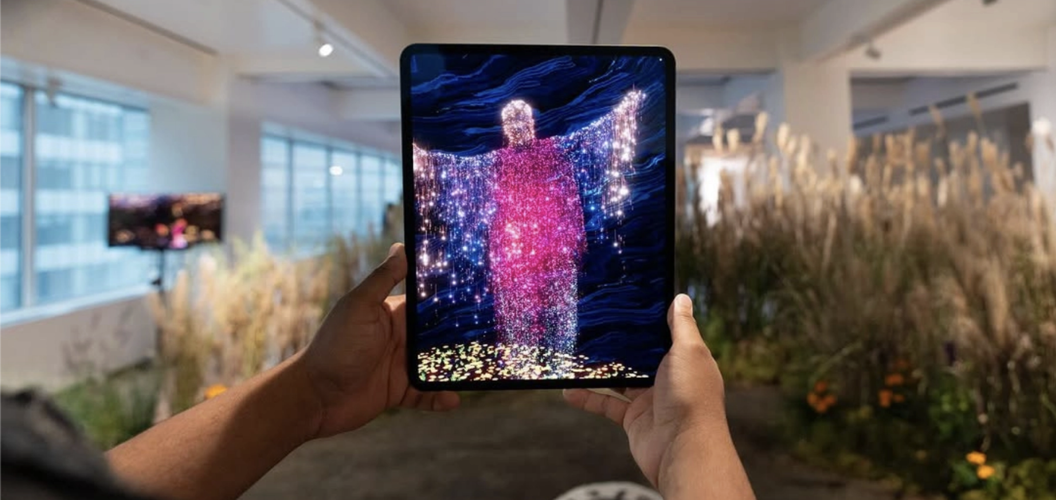 Person holding a tablet with an illuminated digital art display of a colorful, glowing human figure, with background indoor space with large windows and plants