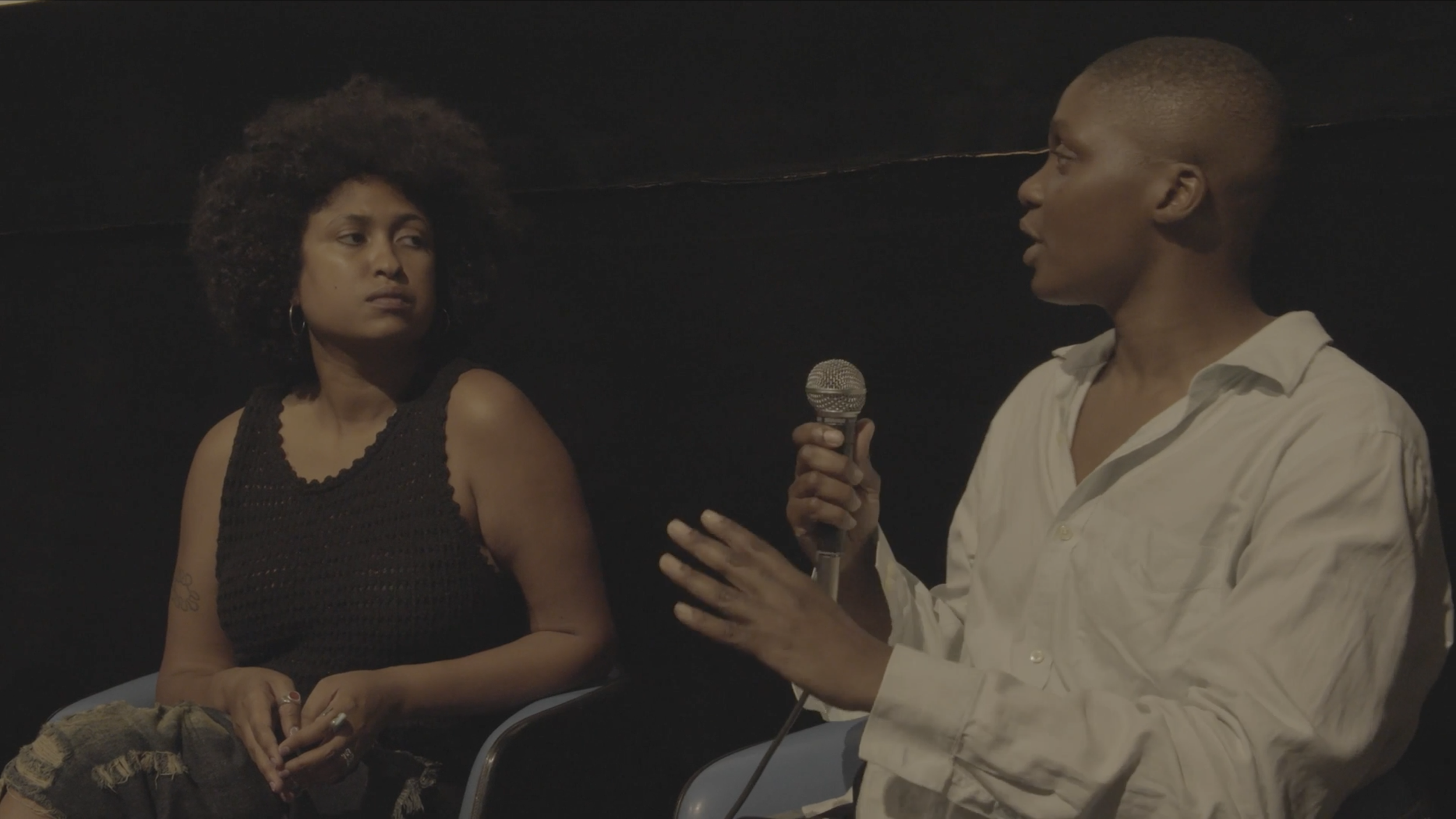 A woman holding a microphone and speaking while a woman listens with a serious expression in a dark room.
